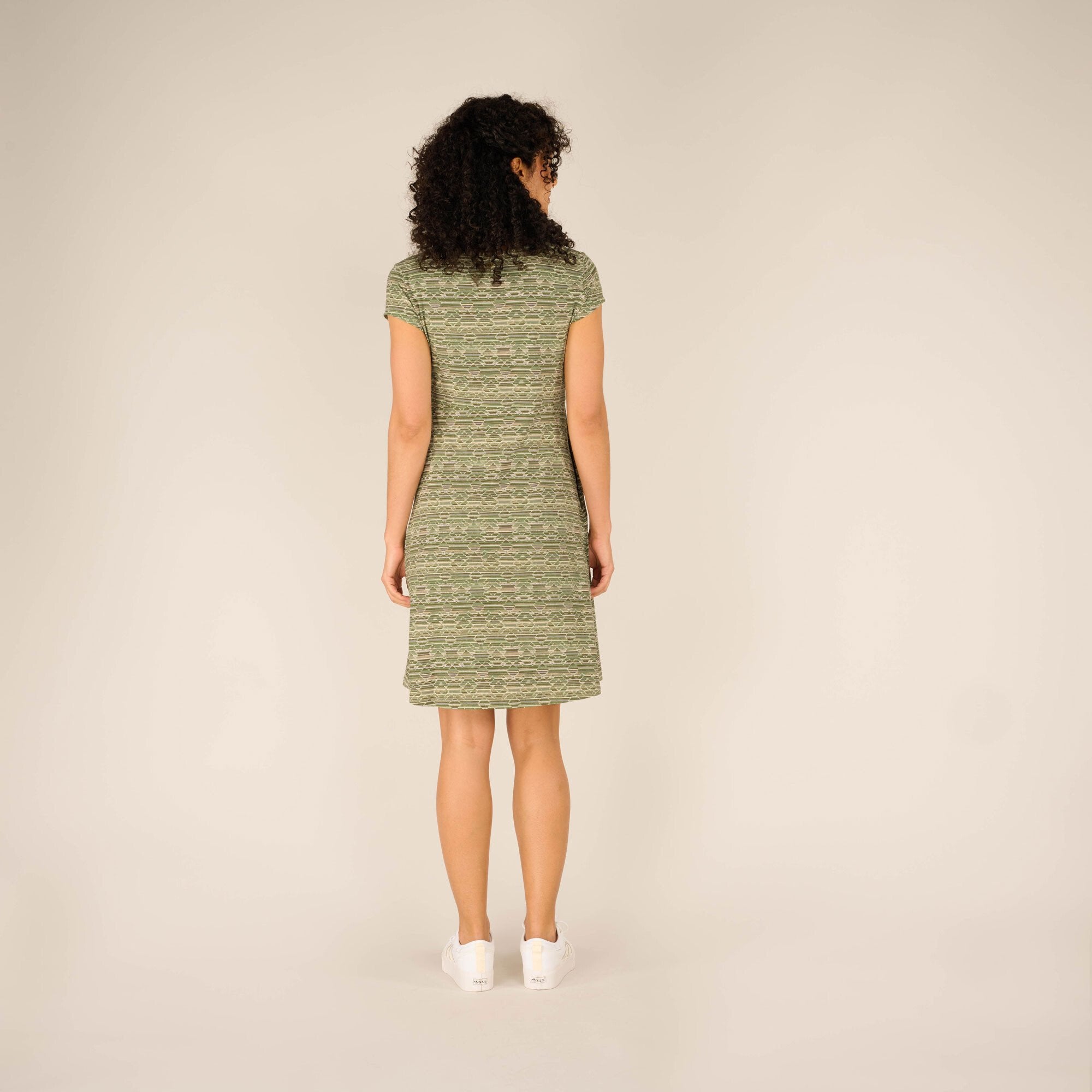 A full rear view of the woman in the Sherpa Adventure Gear Padma Wrap Dress in Green. The dress fits snugly around her upper body and gently flares down to the knees. She is standing straight with her hands relaxed by her sides, wearing clean white trainers.