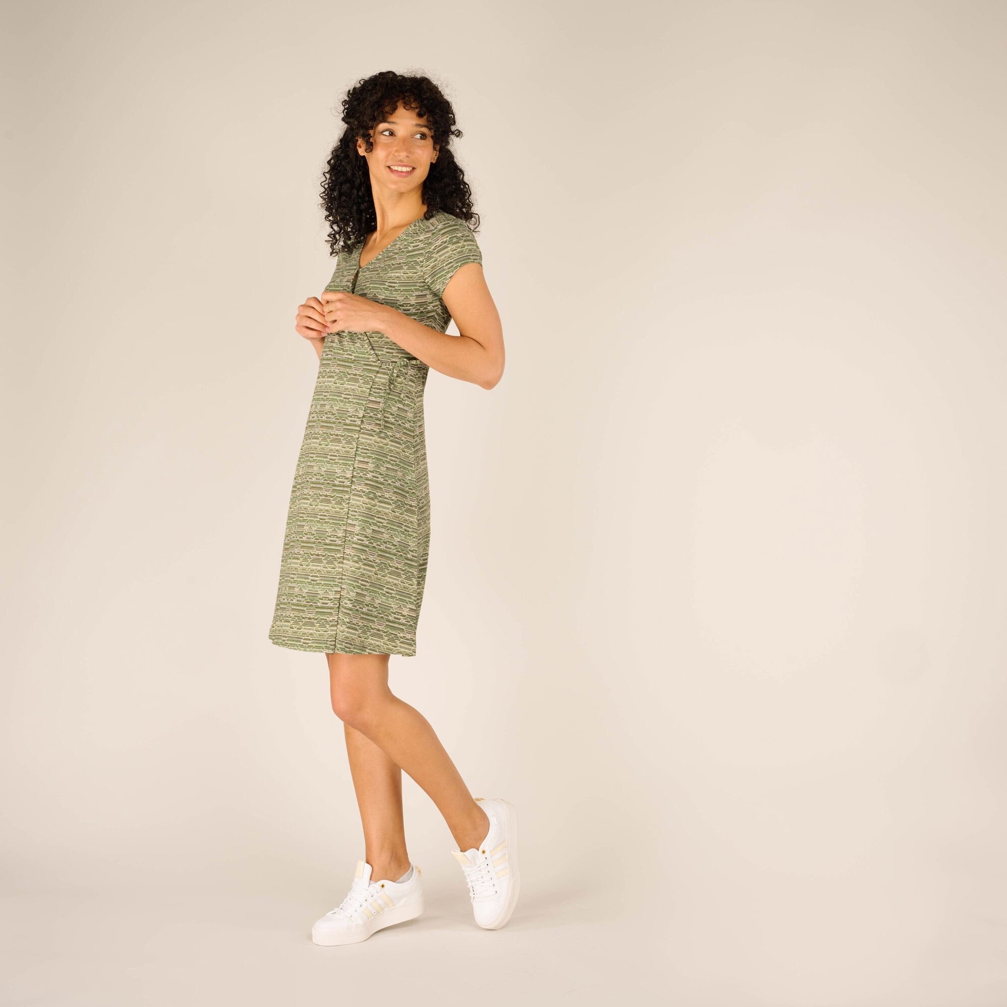 A full-body side view of the woman modeling the Sherpa Adventure Gear Padma Wrap Dress in Green. She stands slightly at an angle, holding her hands together in front of her. The dress’s wrap design and drape are visible, along with her relaxed, smiling pose.