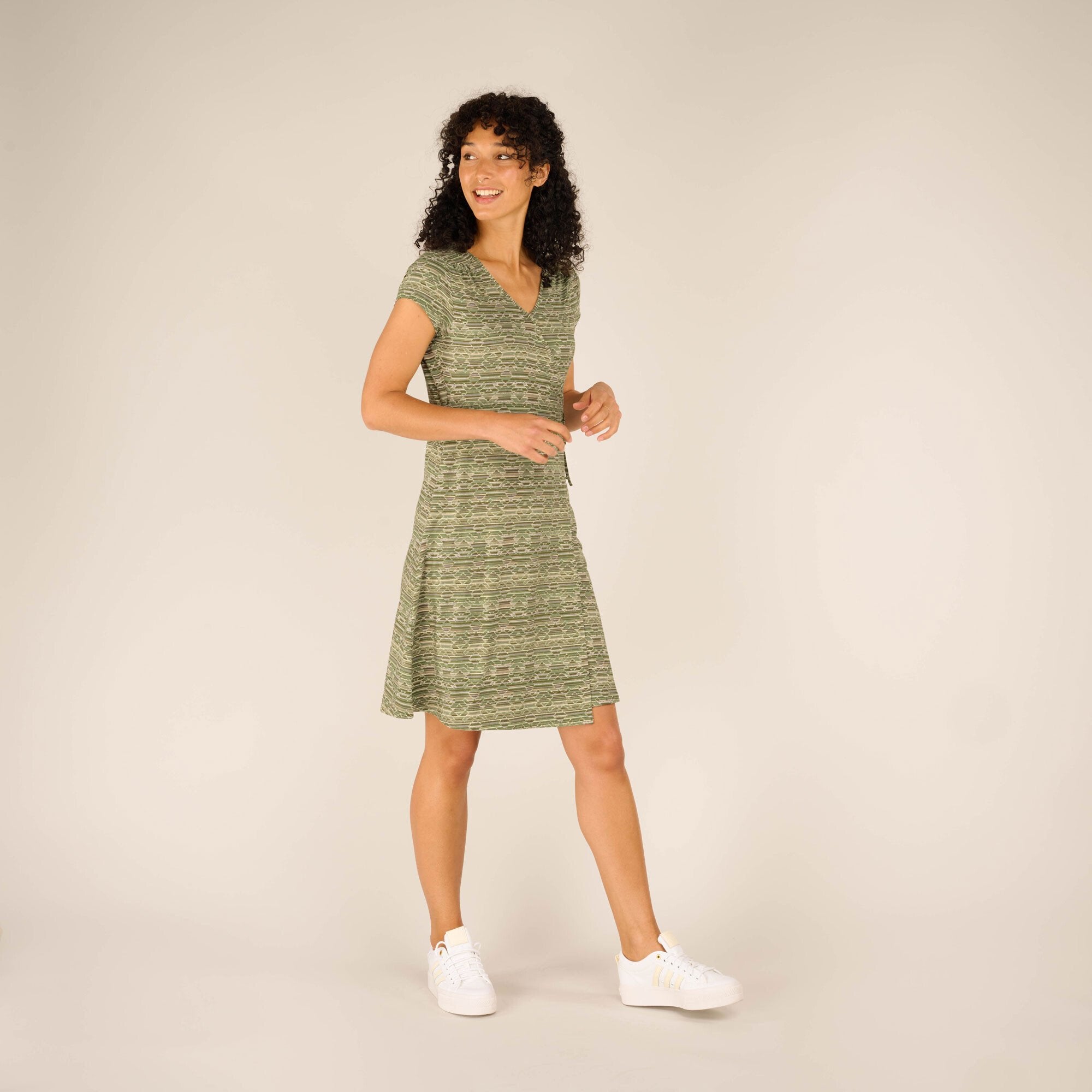 A three-quarter view of the woman, showcasing the Sherpa Adventure Gear Padma Wrap Dress in Green’s flattering silhouette. She stands with one hand lightly resting by her side while looking to her left with a cheerful expression. The short sleeves, V-neckline, and knee-length hemline of the dress are visible, along with her white trainers, which complement the relaxed vibe.