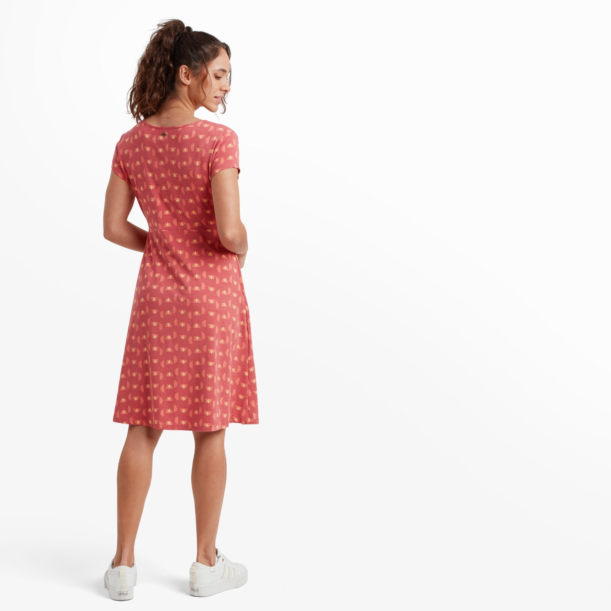 The back view of the woman in the Sherpa Adventure Gear Padma Wrap Dress in Red. The dress flows down to knee length, with the geometric print continuing throughout. Her posture highlights the subtle A-line shape of the dress.