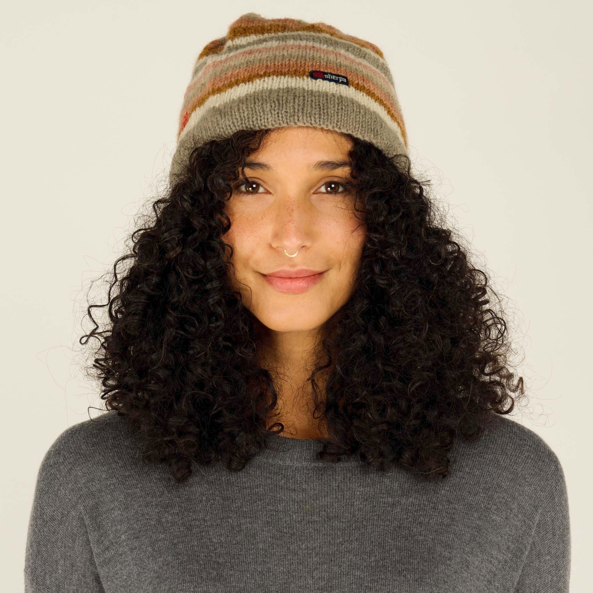 A woman with voluminous, curly black hair wears a Sherpa Adventure Gear Pangdey Hat in Grey with horizontal stripes in shades of beige, brown, and muted orange. The beanie has a small rectangular "Sherpa" brand label on the front. She has a gentle smile, warm brown eyes, and a gold septum ring. She is also wearing a dark gray sweater. The background is a soft, neutral shade.