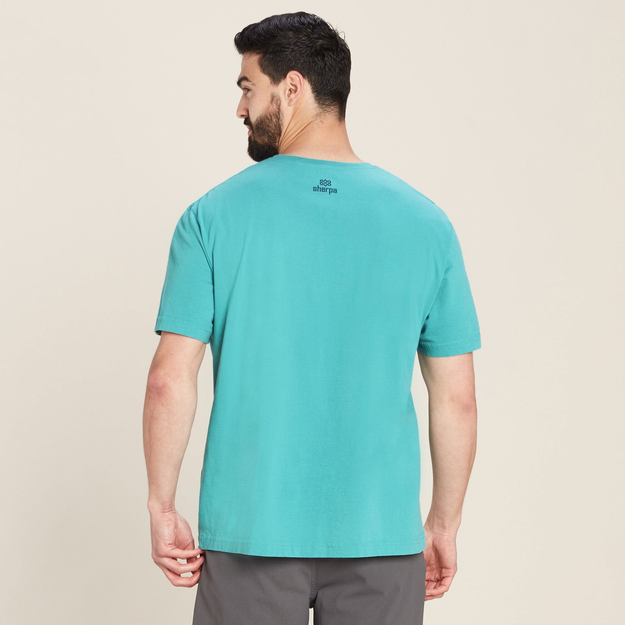 The same man is shown from the back, wearing the Sherpa Adventure Gear Parsa T-Shirt in Blue. On the upper back, just below the neckline, the t-shirt has the "Sherpa" logo printed in black. The t-shirt fits comfortably across his shoulders, and the sleeves fall just above the elbow. His hands are relaxed at his sides.