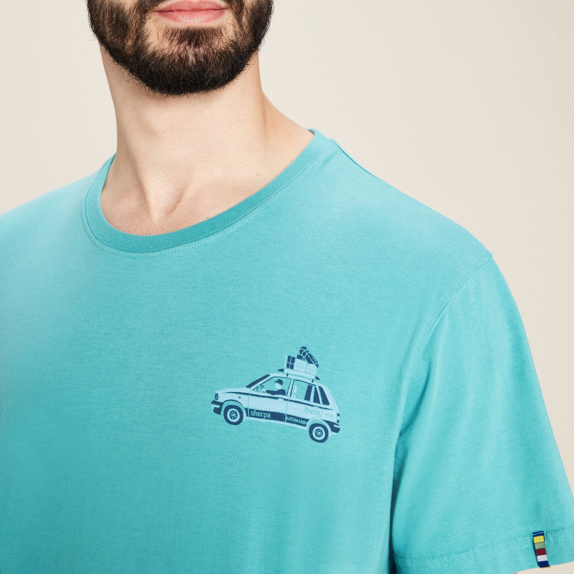 A close-up shot of the Sherpa Adventure Gear Parsa T-Shirt in Blue's chest graphic, showing a detailed print of a car loaded with luggage on the roof. The car has text reading "Sherpa" and "Kathmandu" on the side. The t-shirt’s fabric is smooth, and the neckline stitching is visible. The model’s beard and the lower part of his face can be seen in the frame.