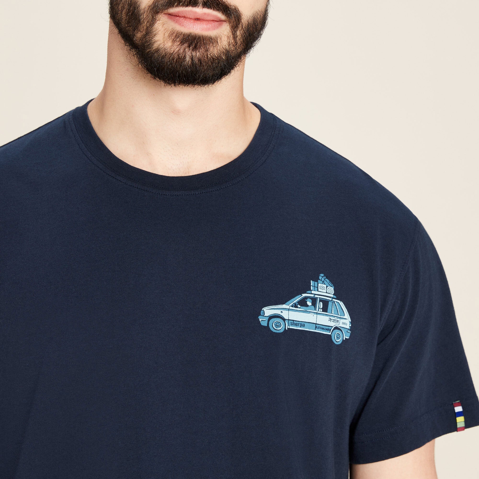 Close-up detail shot of the upper chest area of the Sherpa Adventure Gear Parsa T-Shirt in Blue, highlighting the small car graphic in light blue. The graphic depicts a vehicle loaded with luggage on the roof, with the word "Sherpa" underneath. The image also shows the t-shirt's ribbed neckline and soft fabric texture. A small woven trim with the Sherpa prayer flag colors is sewn into the left sleeve hem.