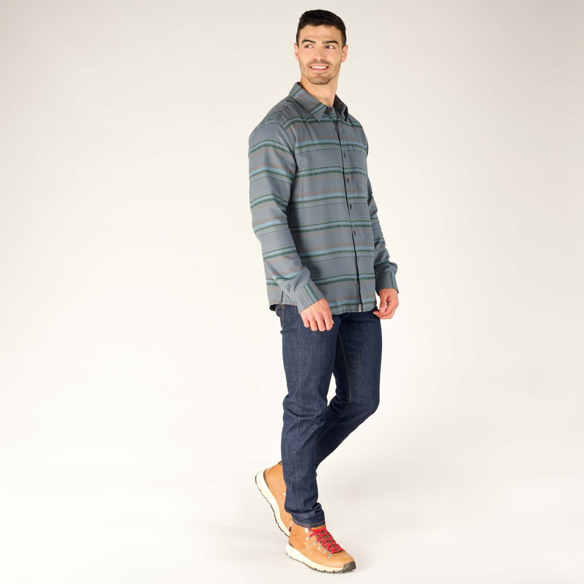 The same man is shown in a side-angled stance, showcasing the full length of the Sherpa Adventure Gear Pasang Shirt in Blue, jeans, and boots. His left hand is casually resting at his side while he looks slightly off-camera, creating a natural and dynamic pose.