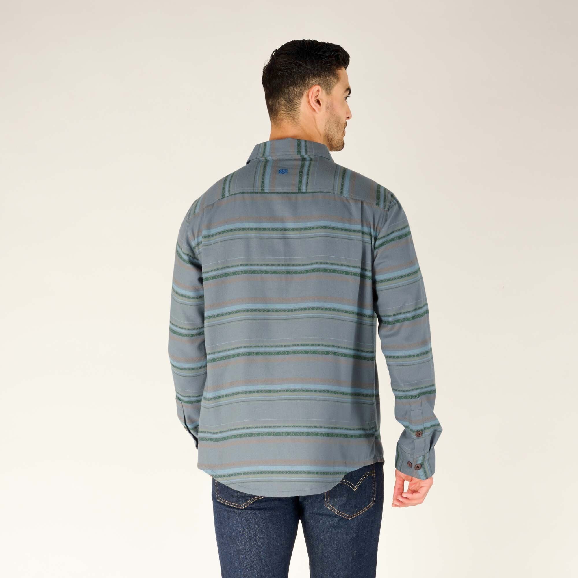 The man is standing with his back to the camera, highlighting the back design of the Sherpa Adventure Gear Pasang Shirt in Blue. The striped pattern continues seamlessly, and the shirt's length and fit are showcased along with the blue denim jeans.