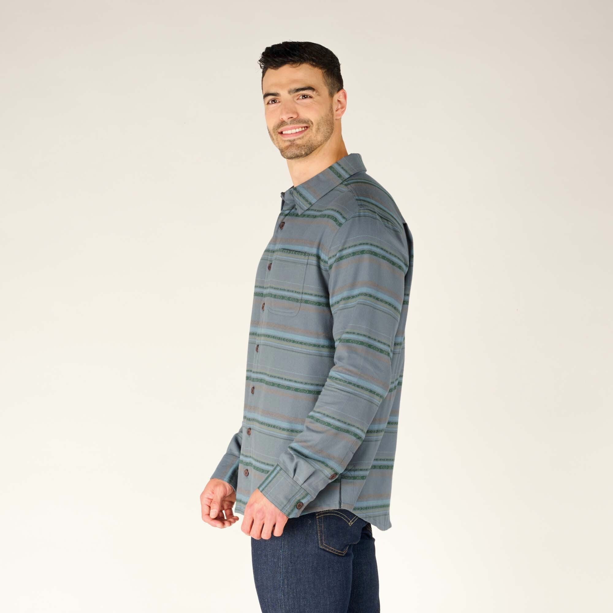 The model is slightly turned to the side, smiling over his shoulder toward the camera. The image captures the casual fit of the Sherpa Adventure Gear Pasang Shirt in Blue and its compatibility with outdoor or casual wear styles.