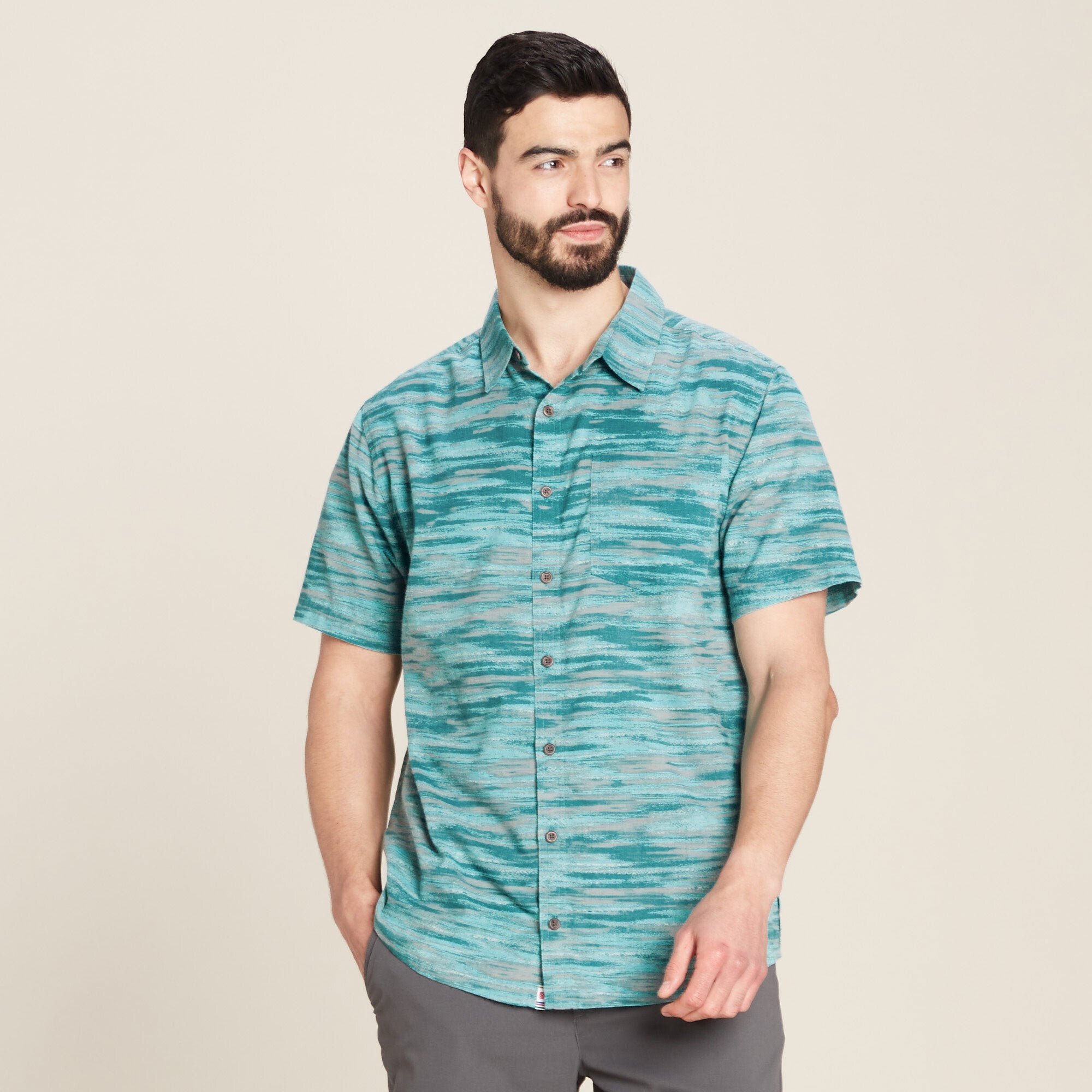 A front-facing view of a man wearing a Sherpa Adventure Gear Pathhar Short Sleeve Shirt in Blue with a horizontal stripe pattern resembling watercolor brushstrokes in shades of teal and gray. The shirt features a relaxed collar, a chest pocket on the left side, and is paired with gray pants. The man has a neat beard and short dark hair, and he is looking to his left with a neutral expression.