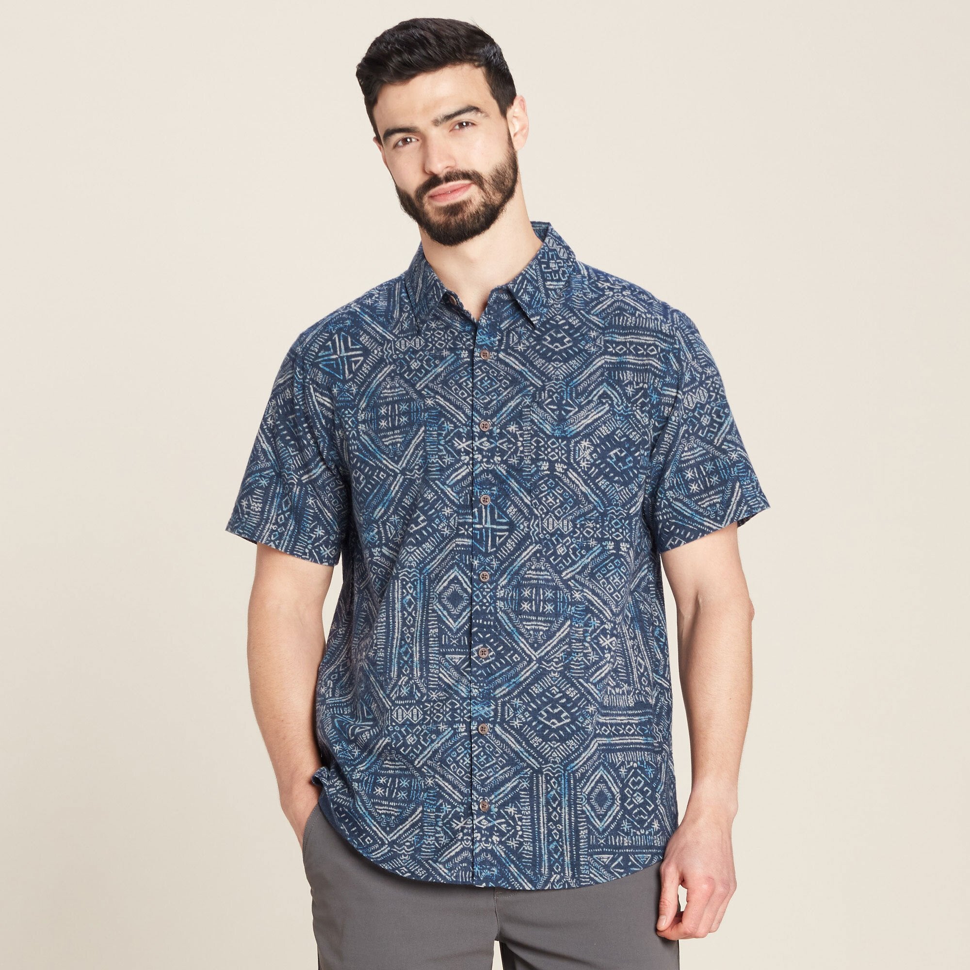 The first image showcases a model wearing a Sherpa Adventure Gear Pathhar Short Sleeve Shirt in Blue in a vibrant blue color with an intricate geometric pattern resembling traditional artwork. The pattern features various shapes and designs in shades of blue and white, adding depth and texture. The shirt has a relaxed fit, a standard collar, and visible buttons down the front. The model stands facing forward with hands in pockets, highlighting the casual yet stylish appeal of the shirt.