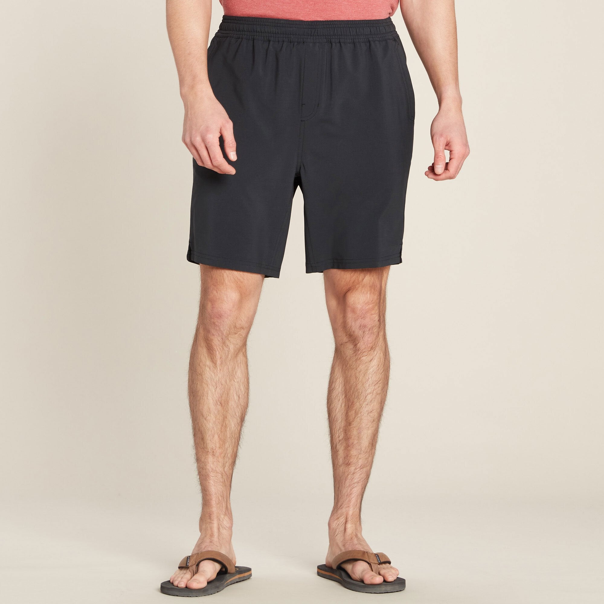 A male model is wearing Sherpa Adventure Gear Pemba Shorts in Black with an elastic waistband. The shorts have a relaxed fit, and the model is also wearing a red t-shirt and brown flip-flops. The background is a neutral beige color.