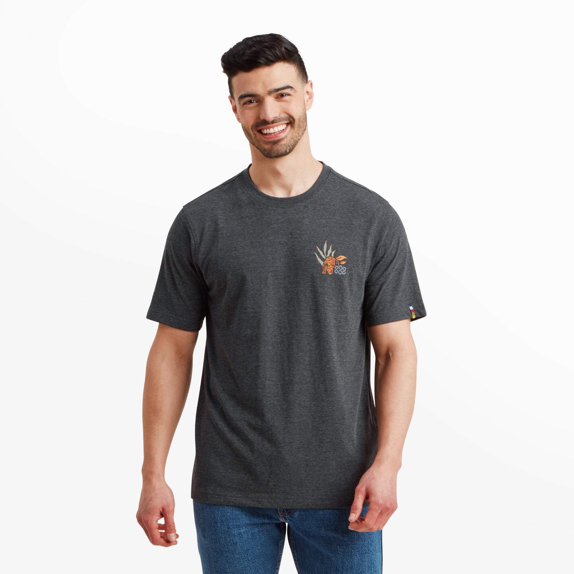 A smiling male model wearing a Sherpa Adventure Gear Purpose T-Shirt in Black. The T-shirt has a minimal design featuring a small colorful graphic of a tiger with leaves and geometric motifs on the left chest. The model pairs the T-shirt with blue jeans against a white background.