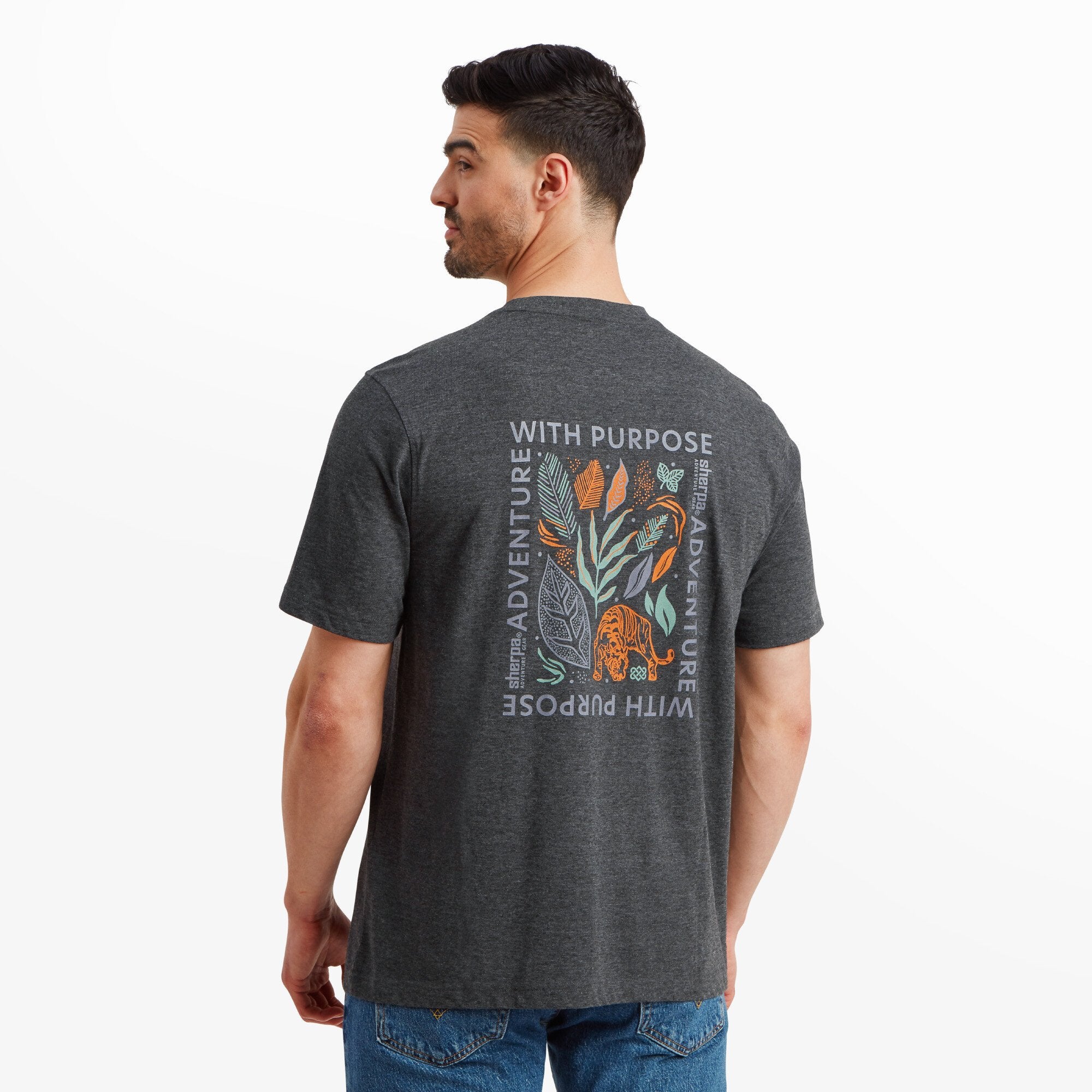 The model facing away from the camera to highlight the back of the Sherpa Adventure Gear Purpose T-Shirt in Black, which features a large, colorful graphic design. The illustration includes botanical leaves, flowers, and a tiger, with text reading "With Purpose" and "Adventure" framing the design in white.