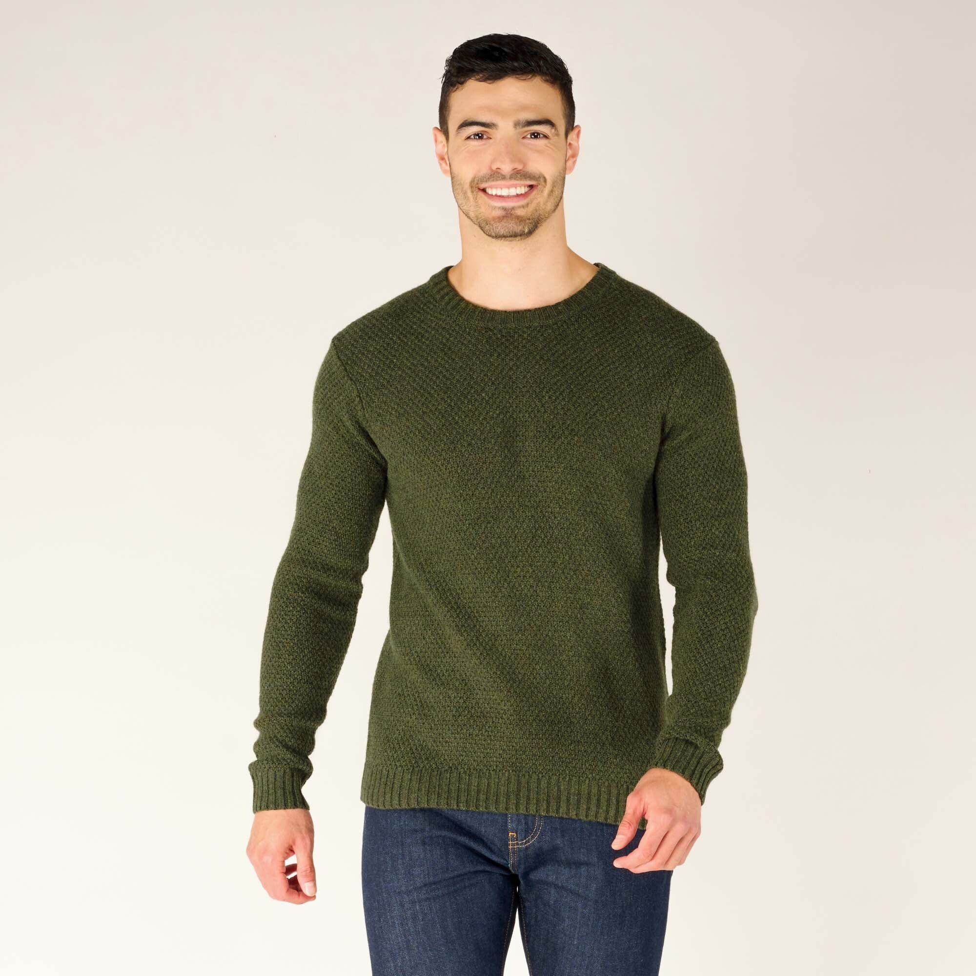 A male model with light skin, short dark hair, and an athletic build stands facing the camera, smiling. He is wearing a Sherpa Adventure Gear Rajen Texture Crew in Green with long sleeves, a ribbed neckline, cuffs, and hem. The sweater has a subtle woven pattern throughout. He pairs the sweater with dark blue jeans.