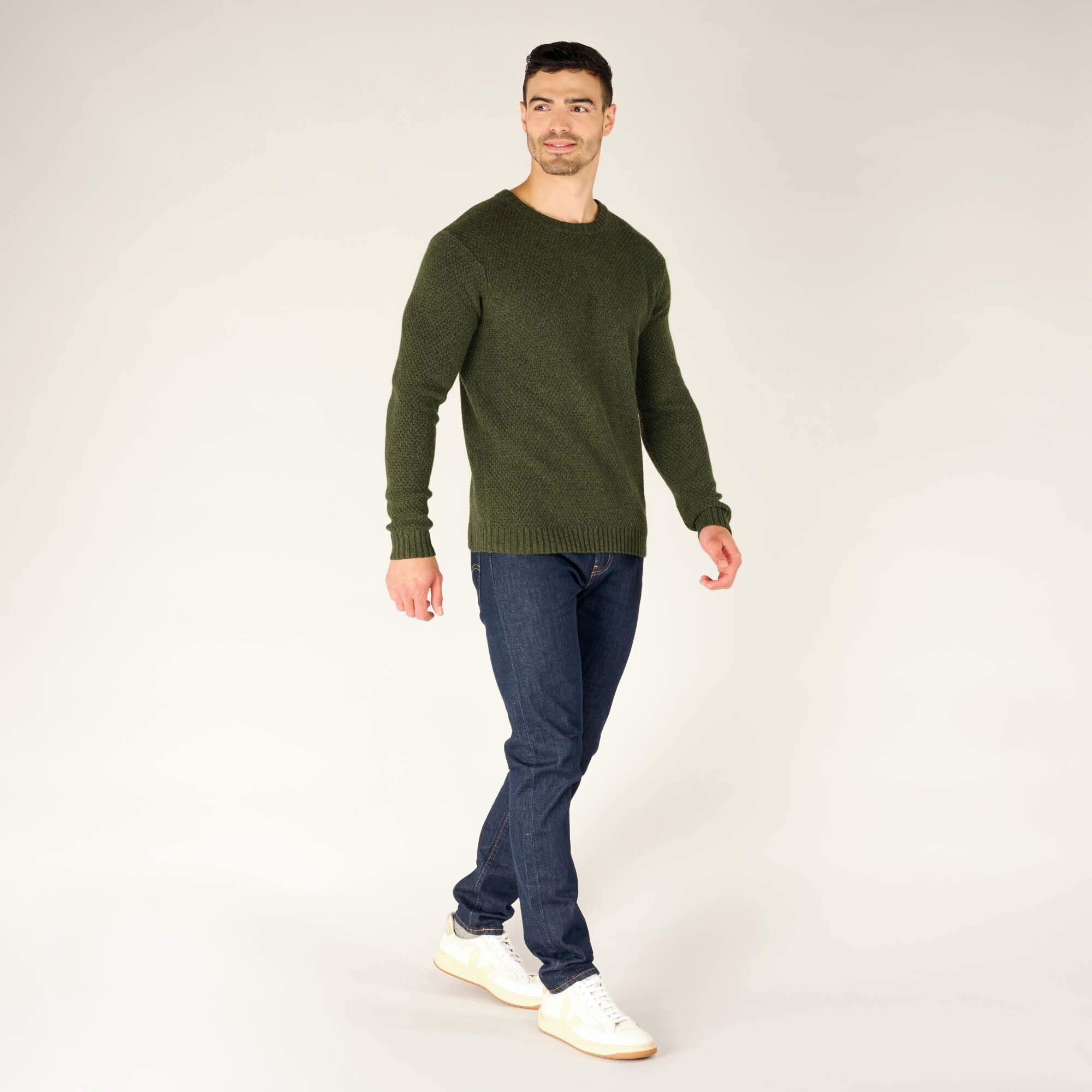 The same male model is captured mid-step, walking slightly to his left while looking off to the side. The green textured crewneck sweater fits comfortably, with the hem falling just below the waist. He is wearing white sneakers with beige accents and dark blue jeans, creating a casual yet polished look.