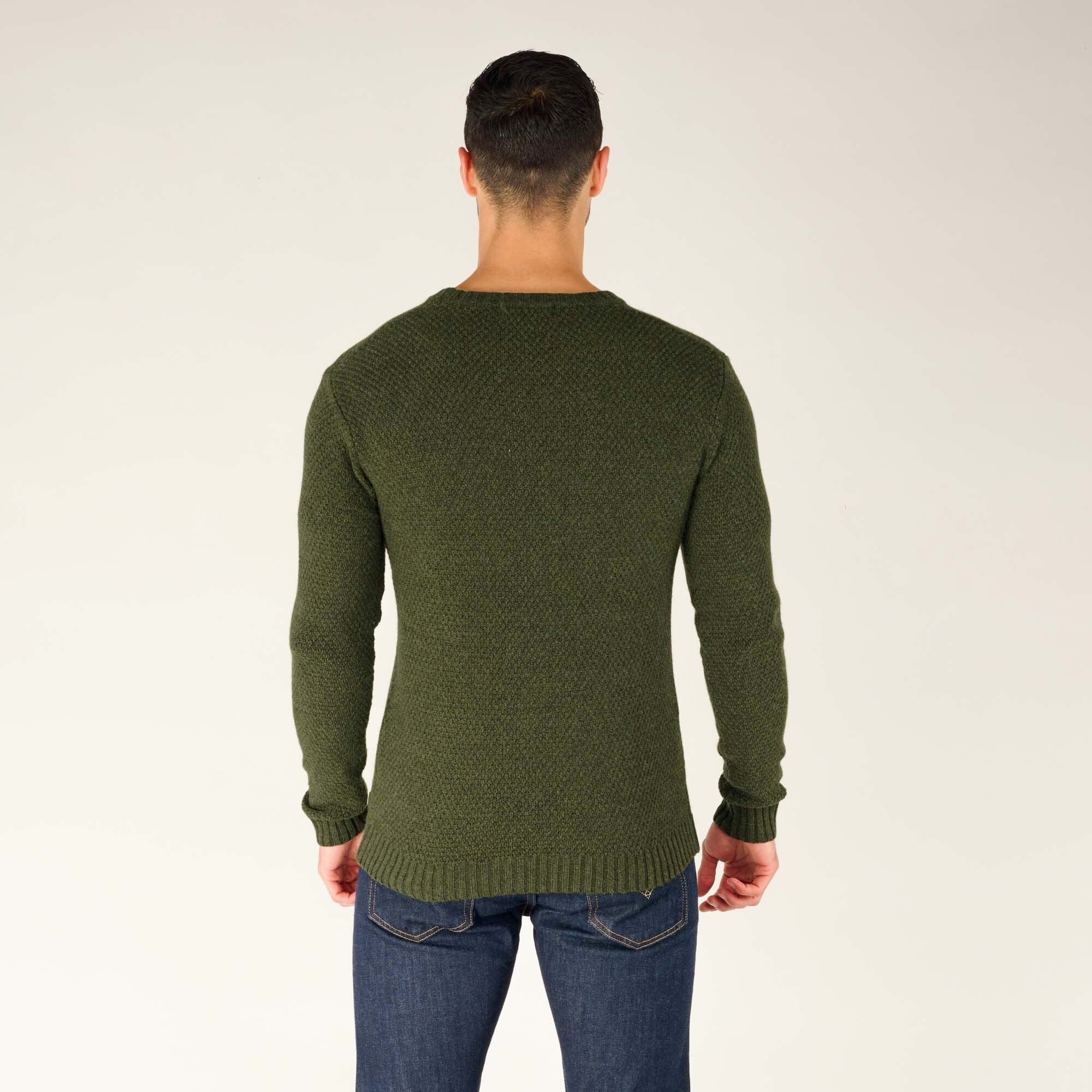 The model faces away from the camera, giving a full back view of the green textured crewneck sweater. The textured knit pattern covers the entire sweater, and the ribbed hem and cuffs ensure a snug fit. The sweater falls just below the waistline, sitting neatly over his dark blue jeans.