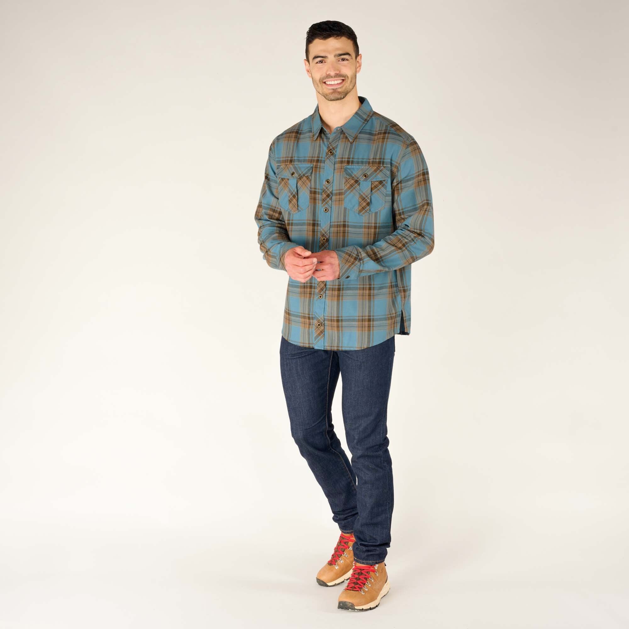 A full-length image of the same male model styled in the Sherpa Adventure Gear Ramoche Shirt in Grey, paired with slim-fit dark blue jeans and tan hiking boots with red laces. He stands in a relaxed, casual pose with hands together.