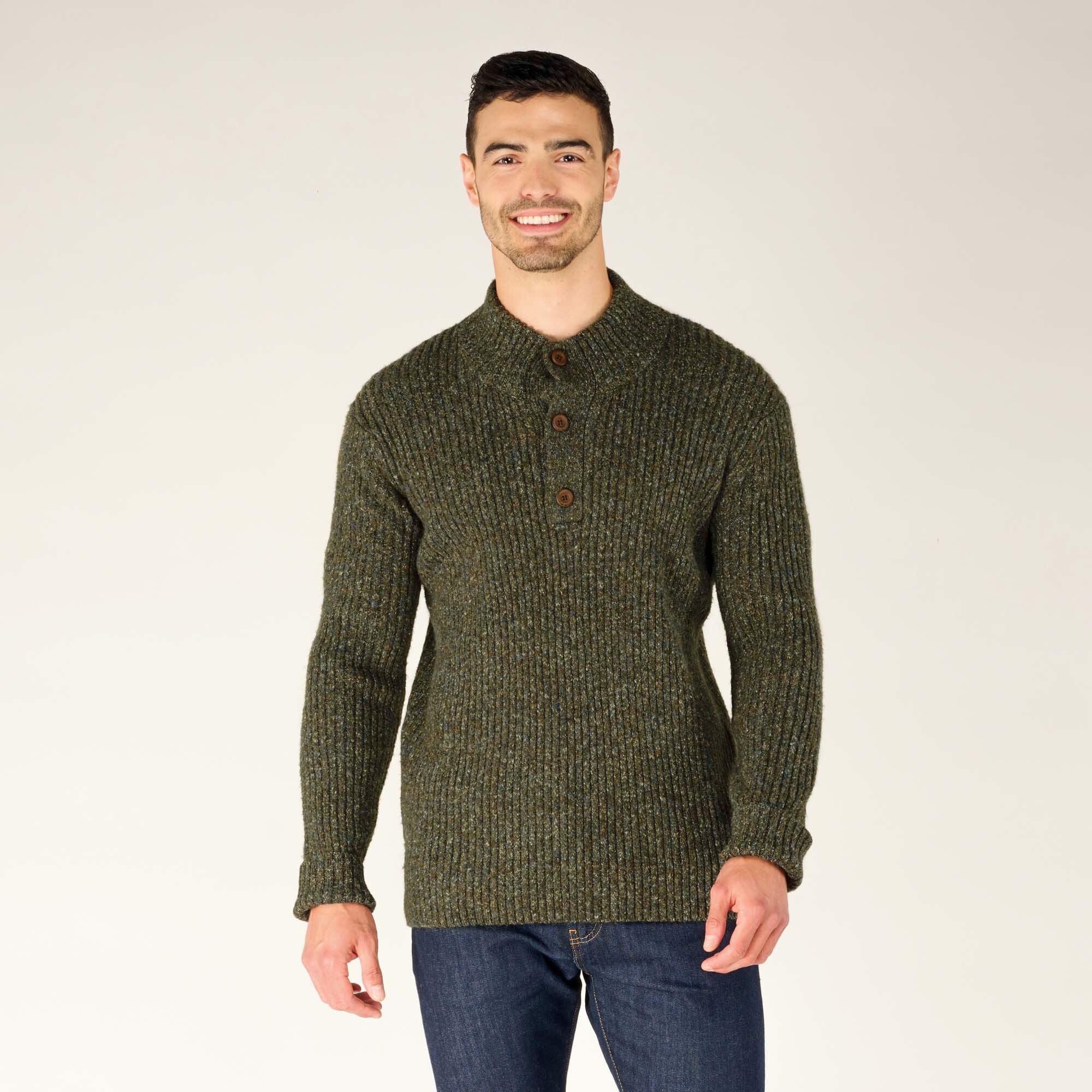 A smiling man is wearing a Sherpa Adventure Gear Ranga Button Mock Neck in Green with a button-up mock neck design. The sweater has a ribbed texture, and he is paired with dark jeans. The photo is set against a neutral beige background.