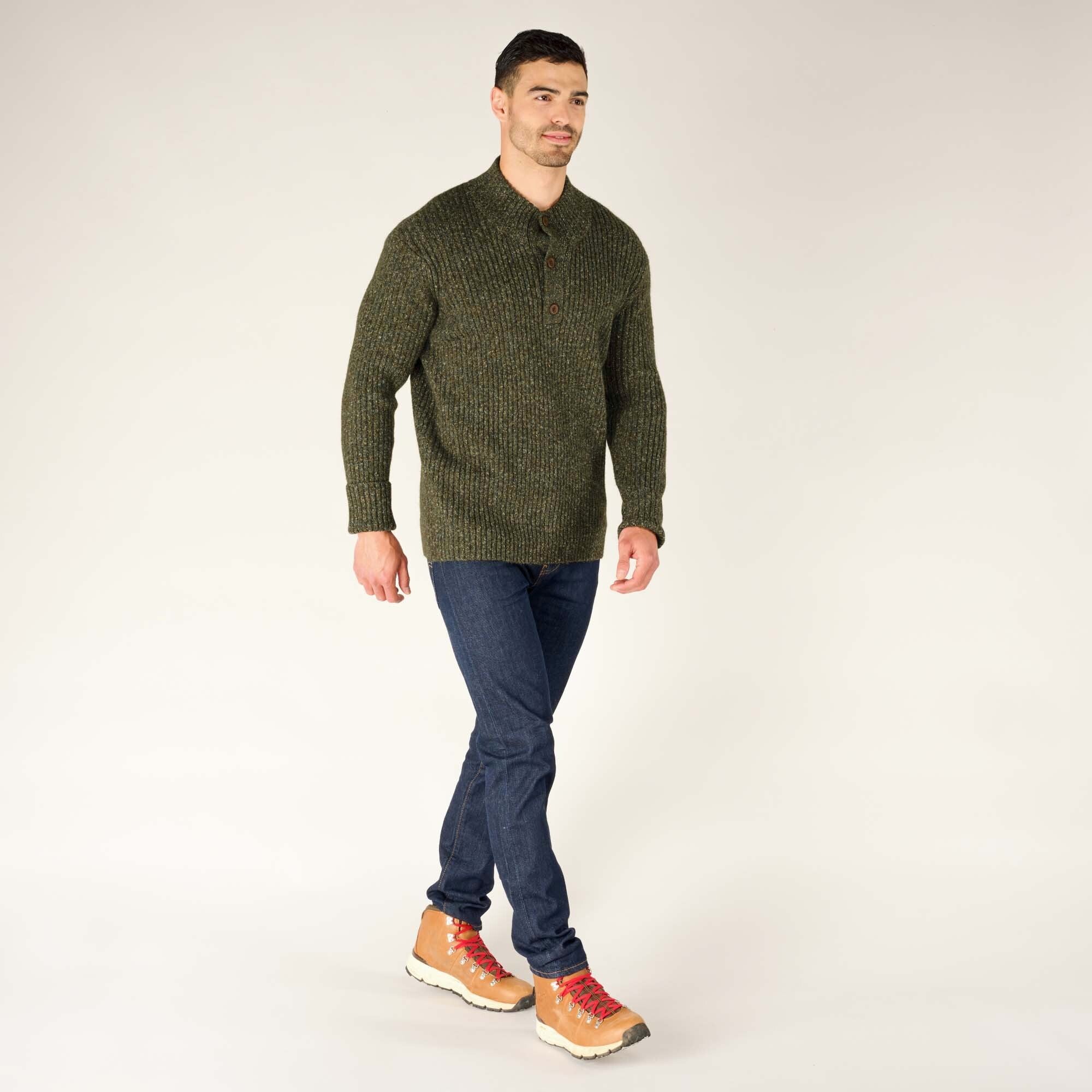 The same man is captured walking confidently while wearing the Sherpa Adventure Gear Ranga Button Mock Neck in Green. He pairs the look with dark jeans and light tan hiking boots featuring red laces. The background is plain and light-colored.