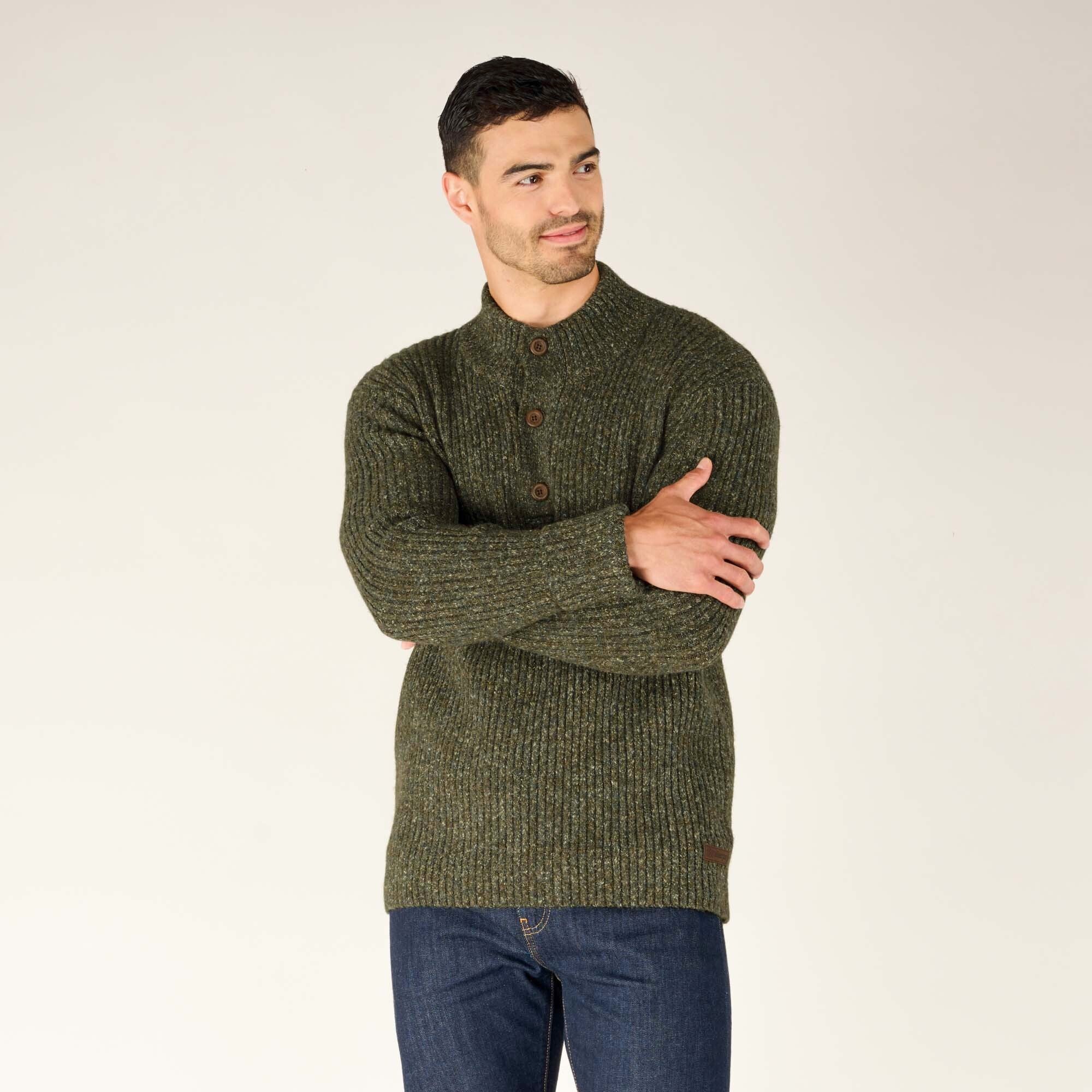 The man stands in a relaxed pose with arms crossed, highlighting the ribbed texture and earthy tones of the Sherpa Adventure Gear Ranga Button Mock Neck in Green. The buttons on the collar are clearly visible, complementing the rugged aesthetic.