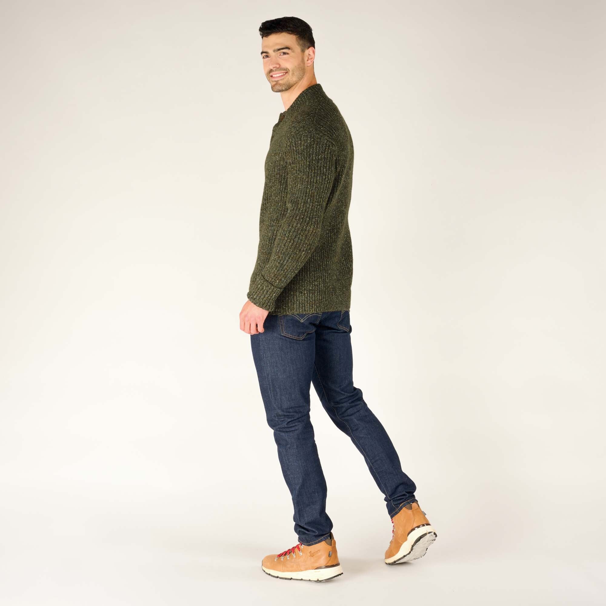 The man turns his body slightly while walking away, highlighting the side profile and back of the Sherpa Adventure Gear Ranga Button Mock Neck in Green. He wears dark jeans and tan hiking boots, making the outfit practical yet stylish.