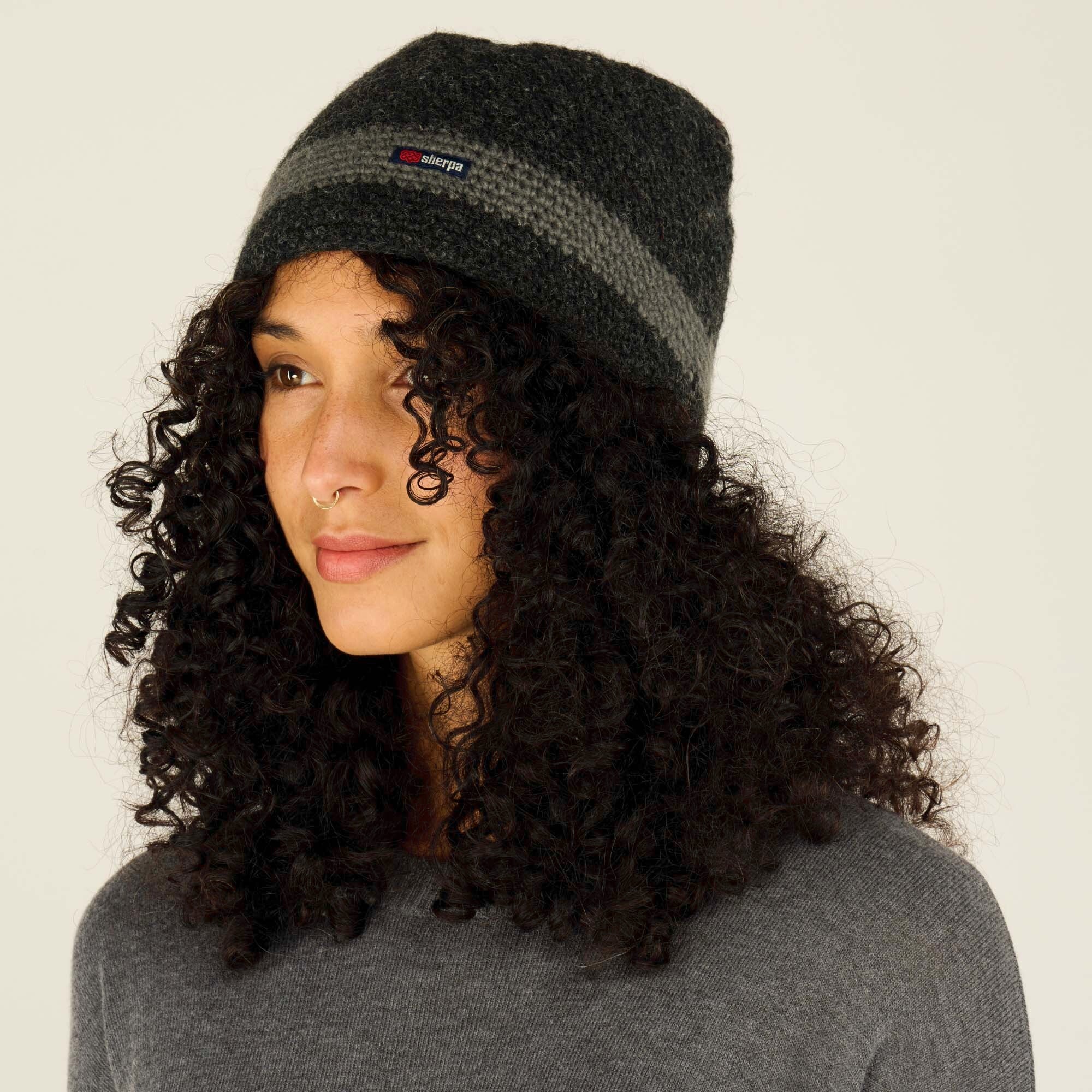 A side-angle view of the same woman wearing the Sherpa Adventure Gear Renzing Hat in Grey with a lighter gray stripe and "Sherpa" logo. The beanie fits snugly, and her curly hair cascades over her shoulders. She gazes off-camera with a calm expression, highlighting the hat's design and texture.