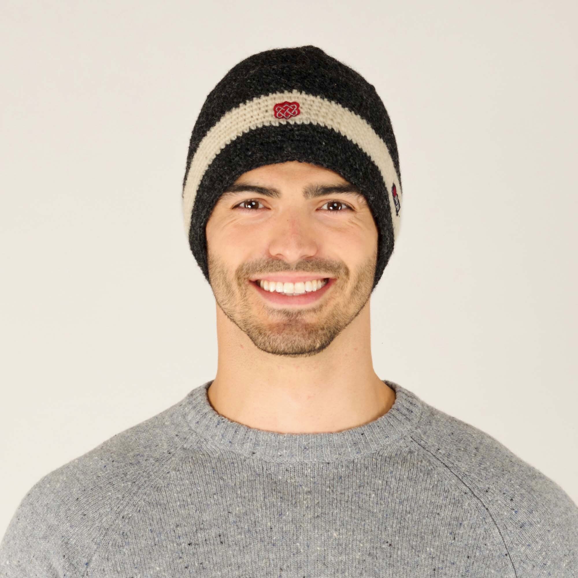 A smiling individual wearing a Sherpa Adventure Gear Renzing Hat in White. The hat features a horizontal cream stripe running across the middle, with a red embroidered symbol at the center. The brand tag with "Sherpa" is stitched on the right side of the cream stripe. The individual is dressed in a grey knit sweater, complementing the cozy aesthetic of the hat.