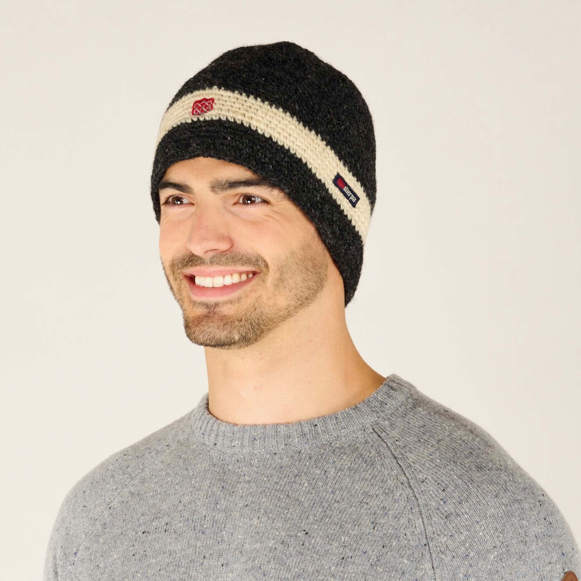 A side profile of the same individual wearing the Sherpa Adventure Gear Renzing Hat in White. The cream stripe and red embroidered symbol are prominently visible, as is the "Sherpa" brand tag stitched on the cream stripe. The texture of the hat's knit pattern is emphasised in this angle, showcasing its detailed craftsmanship. The grey sweater remains visible, enhancing the overall warm and casual look.