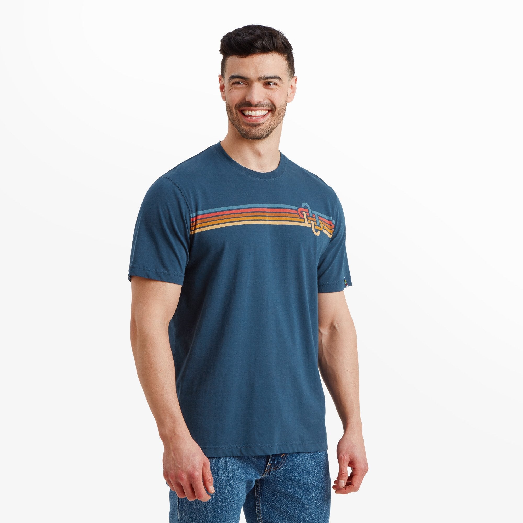 A male model with short dark hair and a beard wears a Sherpa Adventure Gear Retro Knot T-Shirt in Blue. The T-shirt features a horizontal stripe design across the chest with blue, orange, and red stripes, along with an interlocking knot graphic on the upper left side. The model is smiling and standing with his hands relaxed at his sides, wearing blue jeans.