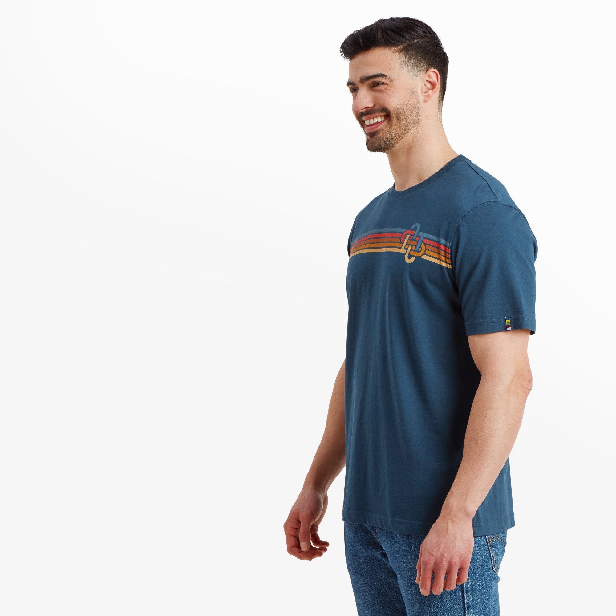 Side view of the same male model wearing the Sherpa Adventure Gear Retro Knot T-Shirt in Blue. The T-shirt's stripe and knot design wraps slightly around the side, and the model is smiling while looking slightly off-camera. He is wearing blue jeans.