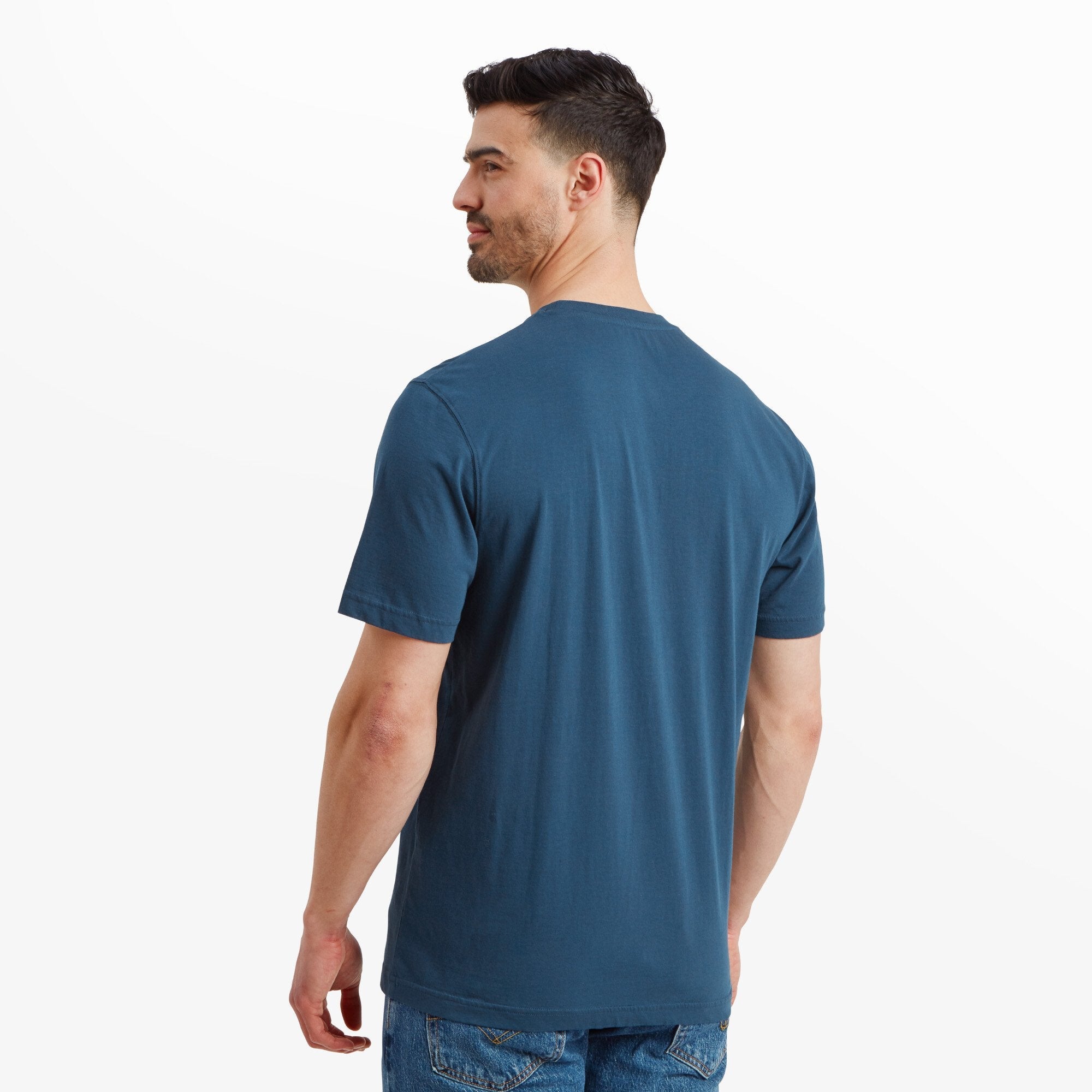 Rear view of the male model showing the back of the Sherpa Adventure Gear Retro Knot T-Shirt in Blue. The back is plain with a solid blue color, and the model’s hands rest naturally at his sides. He is wearing blue jeans.