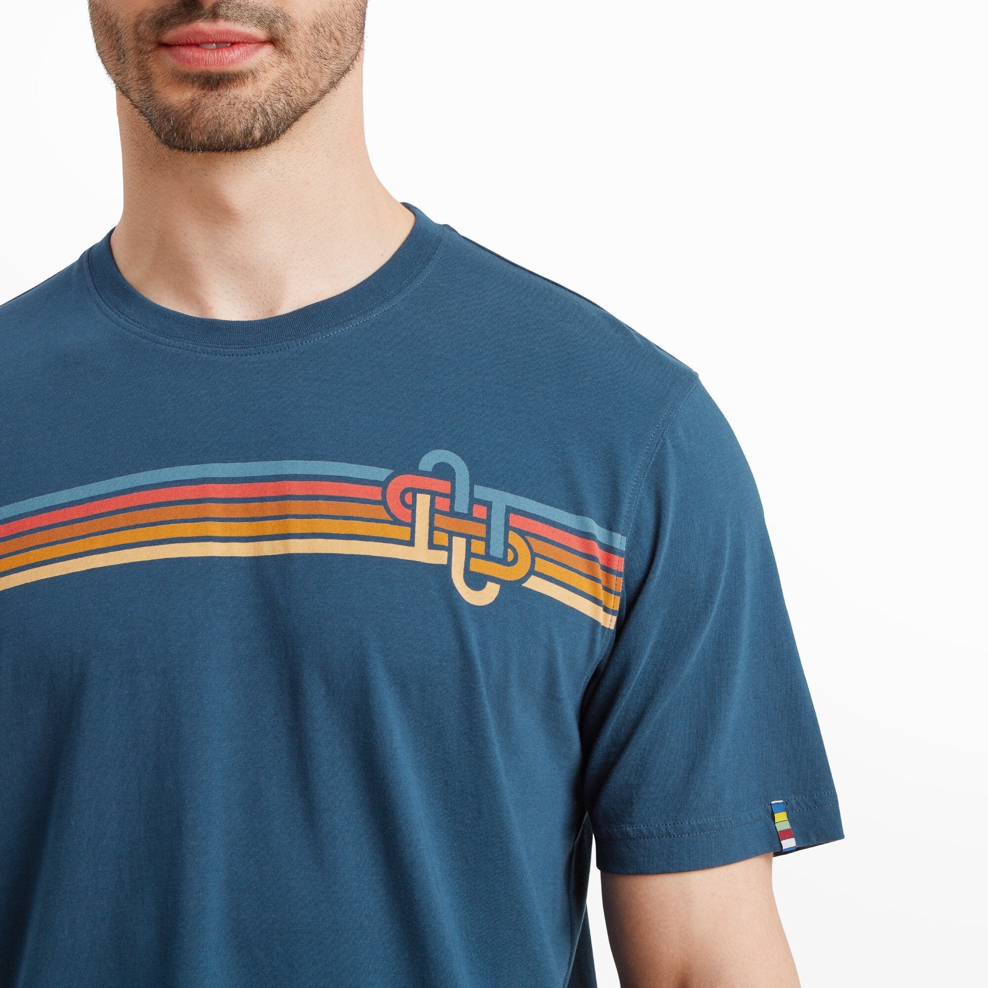 Close-up of the upper chest and shoulder of the Sherpa Adventure Gear Retro Knot T-Shirt in Blue. The focus is on the colorful stripe design and the interlocking knot graphic, with visible details of the fabric texture. A small multicolored woven label is stitched into the left sleeve.