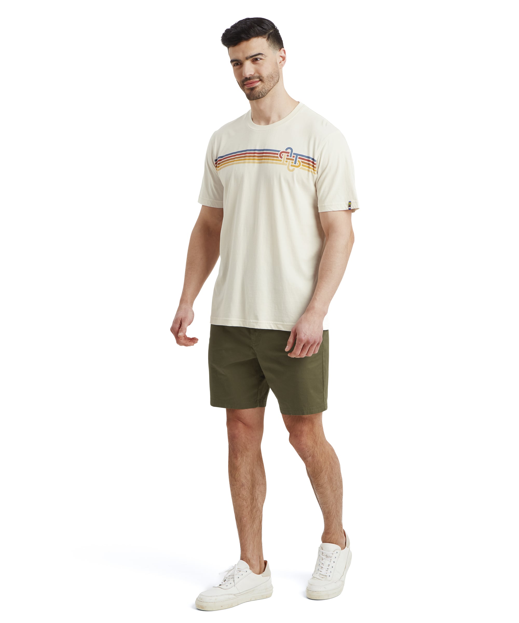 Full-body view of a man dressed in a Sherpa Adventure Gear Retro Knot T-Shirt in White paired with olive-green shorts and white trainers. The man is standing casually with one leg slightly bent, looking off to the side with a smile.