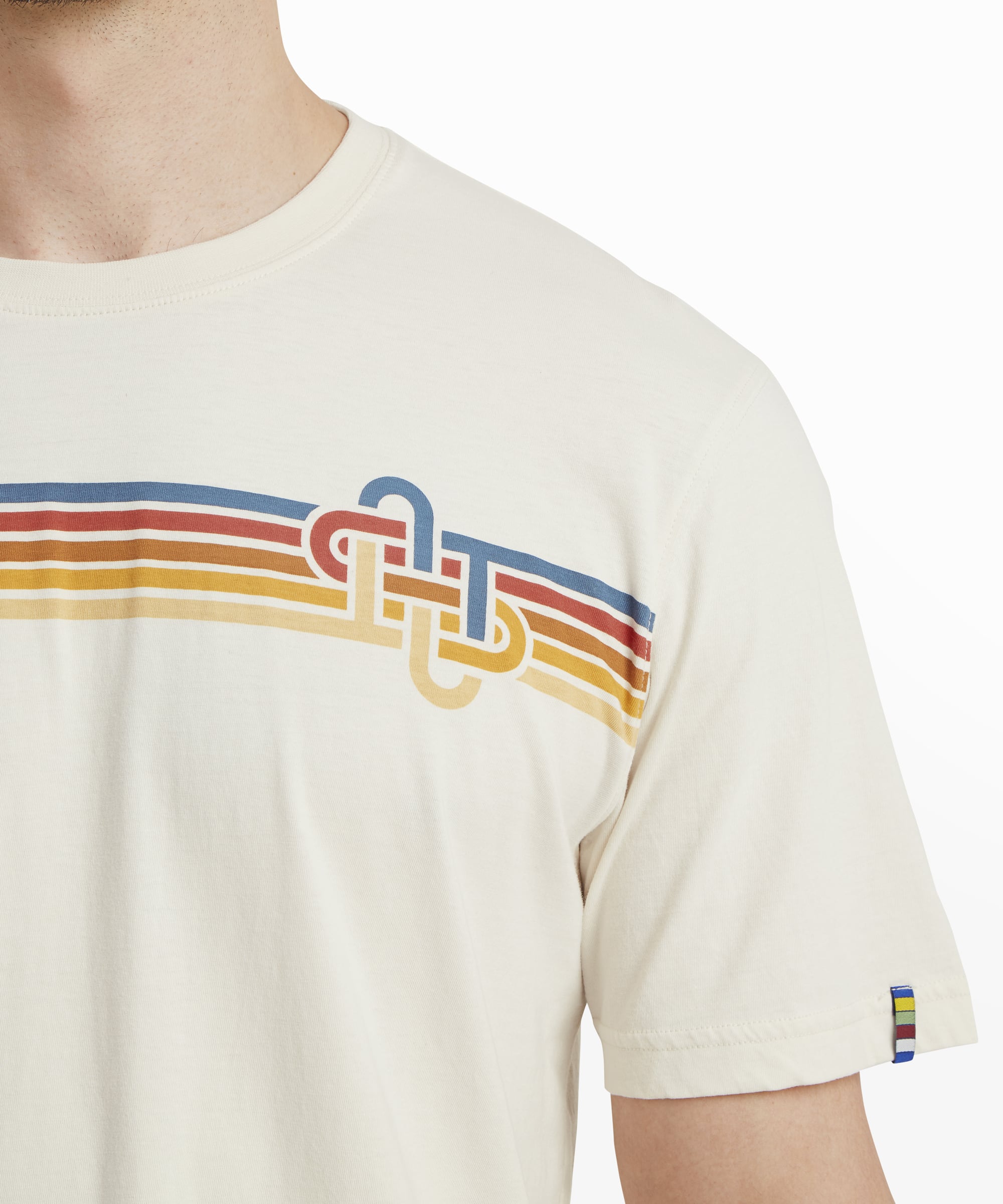 Close-up of the chest area of the Sherpa Adventure Gear Retro Knot T-Shirt in White, highlighting the colorful horizontal stripe design and the knot-inspired logo on the left chest. The sleeve includes a small colorful striped tab detail on the edge, adding a pop of color.