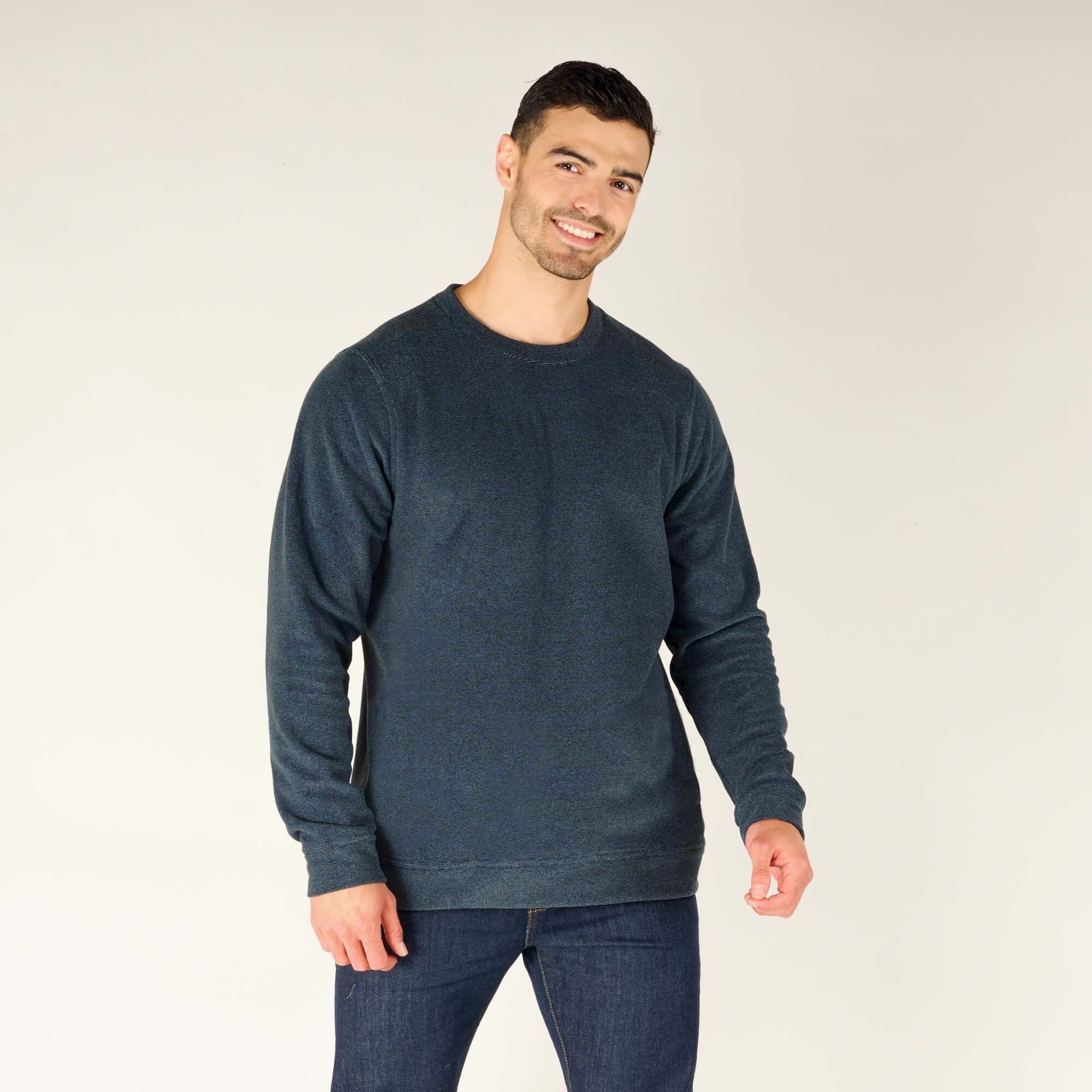 A male model with short dark hair and a beard wears the Sherpa Adventure Gear Rolpa Crew in Blue. The crewneck sweater has a relaxed fit with long sleeves, a soft texture, and subtle stitching along the shoulder seams. The model is standing against a plain white backdrop, smiling slightly, with his arms at his sides. He pairs the sweater with dark blue jeans.