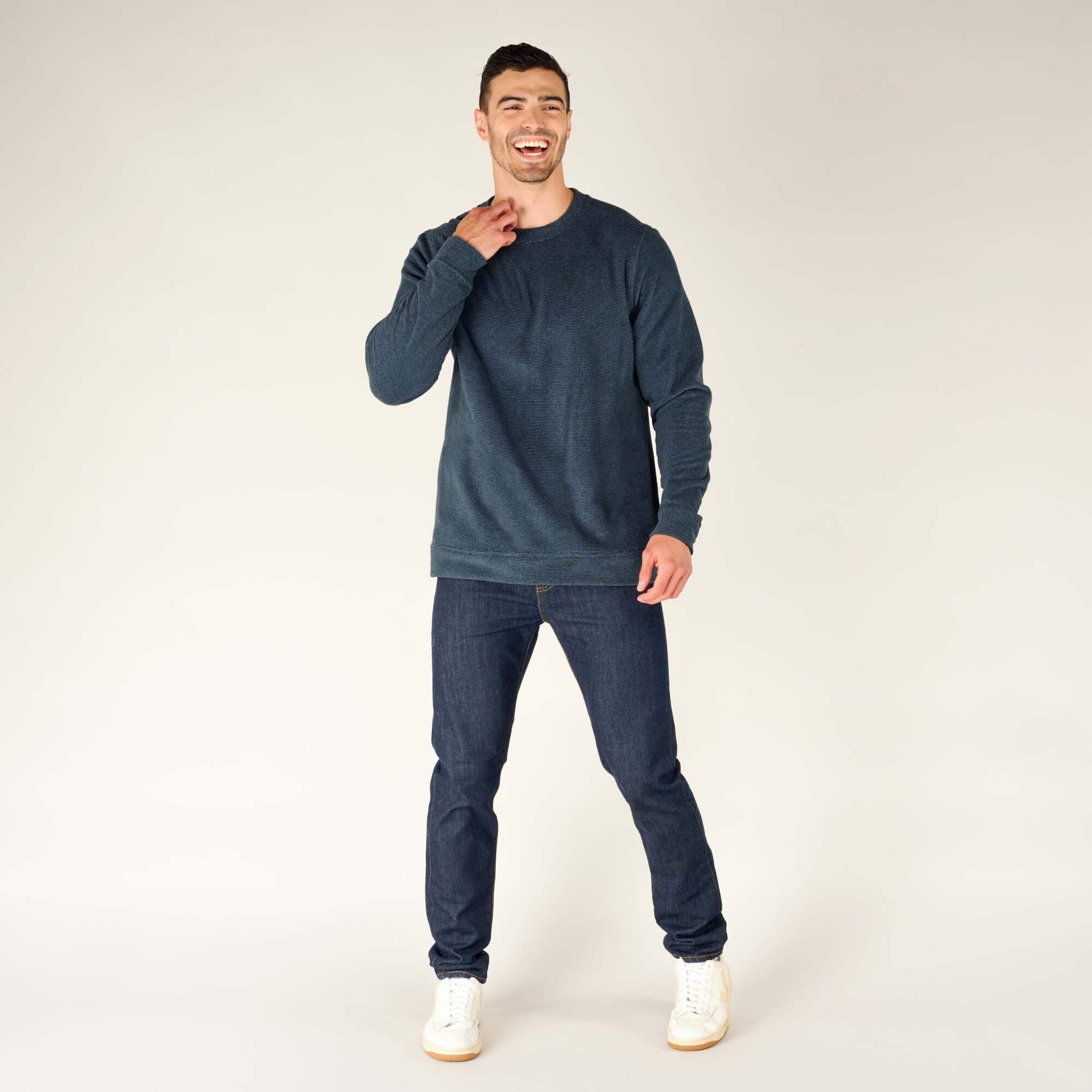 The same male model is captured in a full-body shot wearing the Sherpa Adventure Gear Rolpa Crew in Blue. He is in a relaxed pose, with one hand raised toward his neck as if adjusting the collar and the other hand hanging loosely. The sweater has a straight hem that falls just below the waist. He pairs it with slim dark jeans and white sneakers. The background remains plain white.