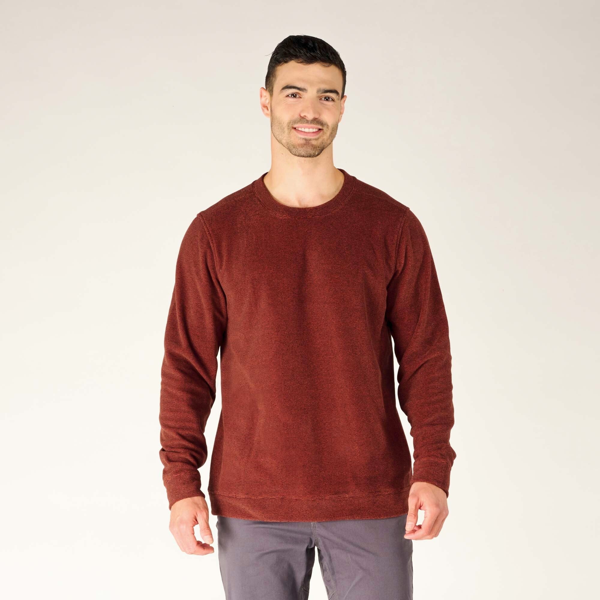 A male model with short dark hair and light skin is shown standing against a neutral beige background, wearing the Sherpa Adventure Gear Rolpa Crew in Red. The long-sleeve crewneck pullover is a rich, earthy red with a subtle heathered texture. The fit is relaxed, with ribbed cuffs and hem. The model smiles gently, with his arms resting naturally at his sides. He pairs the crew with gray pants.