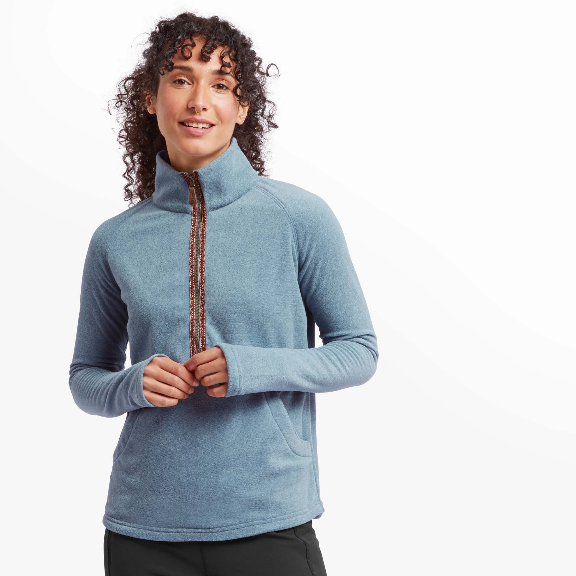 A woman is wearing a Sherpa Adventure Gear Rolpa Eco Half Zip in Blue with a high collar. The fleece features a decorative patterned zipper trim in warm, earthy colors. She stands confidently, smiling gently, with her hands slightly clasped in front of her. The cozy fabric and tailored fit emphasise both comfort and style.