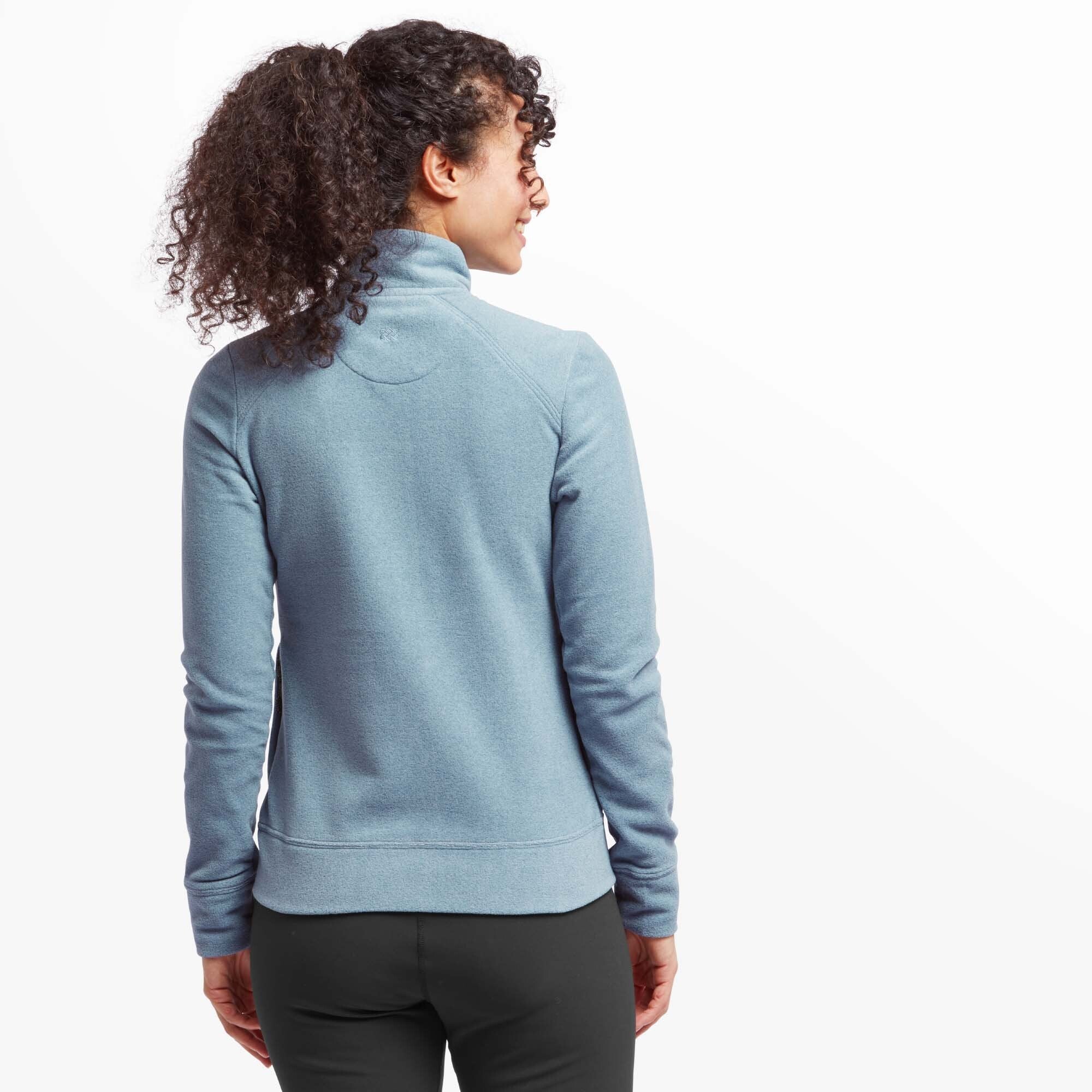 A rear view of the woman wearing the Sherpa Adventure Gear Rolpa Eco Jacket in Blue. Her curly hair is tied back in a ponytail, revealing the back design of the jacket, which has a slightly fitted shape and a subtle curved seam across the upper back. She is turned slightly to the side, smiling. The background is plain white.