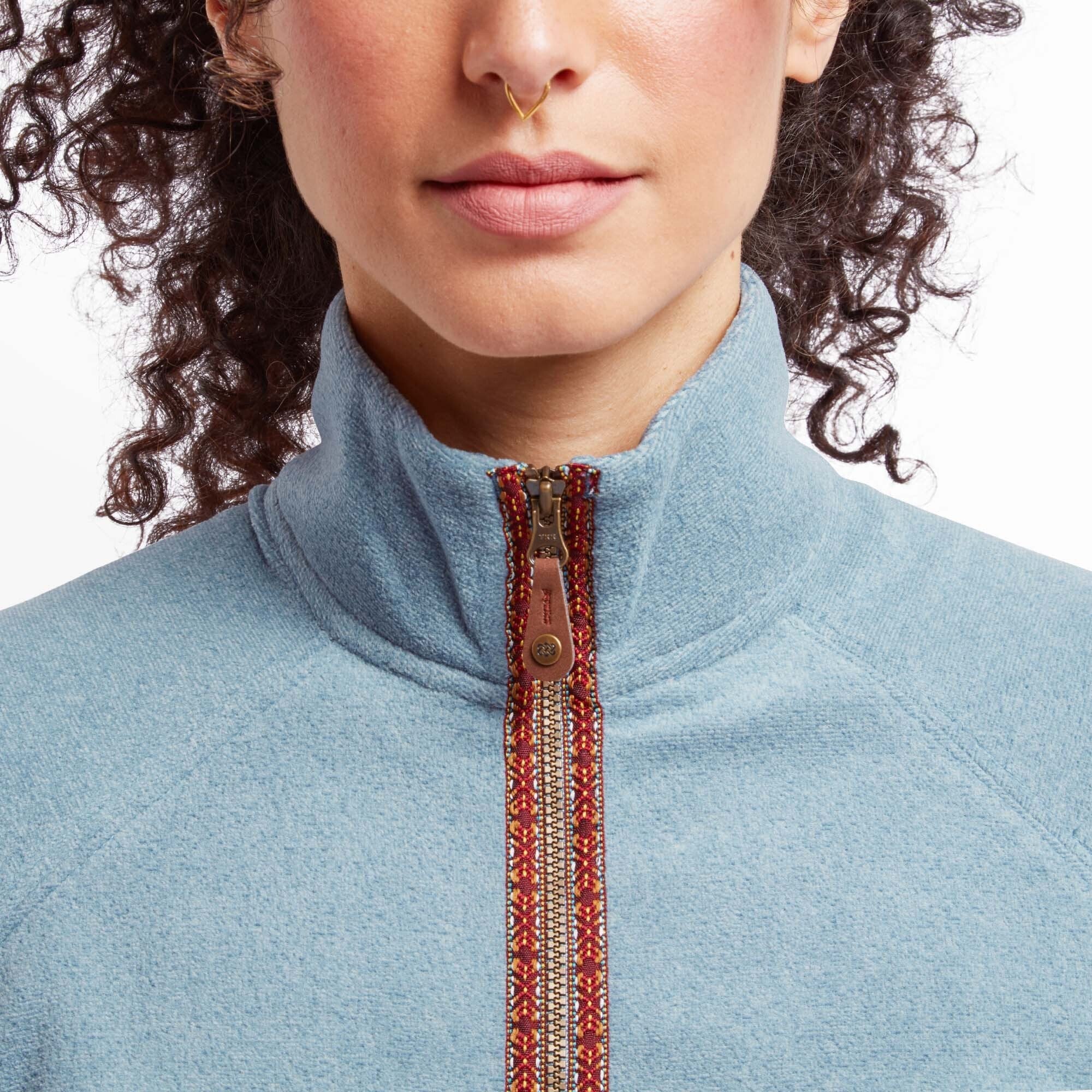 A close-up of the upper part of the Sherpa Adventure Gear Rolpa Eco Jacket in Blue, focusing on the intricate patterned trim along the zipper. The trim features a mix of warm tones, including red, orange, and beige. The woman’s face is partially visible, showing her lips and nose, adorned with a small gold septum ring. The background is plain white.