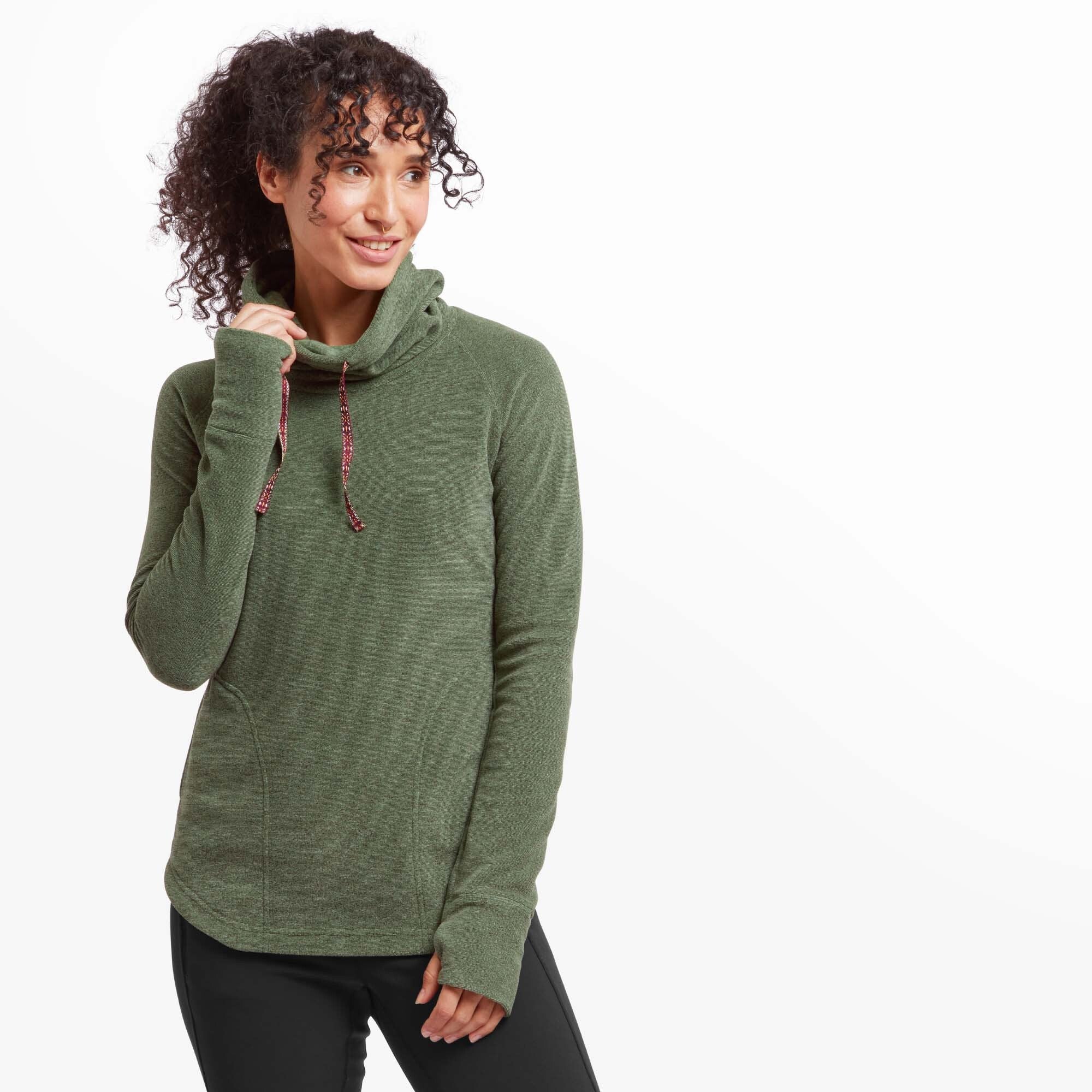 A woman with curly dark hair and a nose ring poses wearing a Sherpa Adventure Gear Rolpa Eco Pullover in Green with a relaxed cowl neck. The pullover has a soft heathered texture and features a contrasting red and white patterned drawstring. She is holding the edge of the cowl neck with one hand and smiling slightly, looking off to the side.