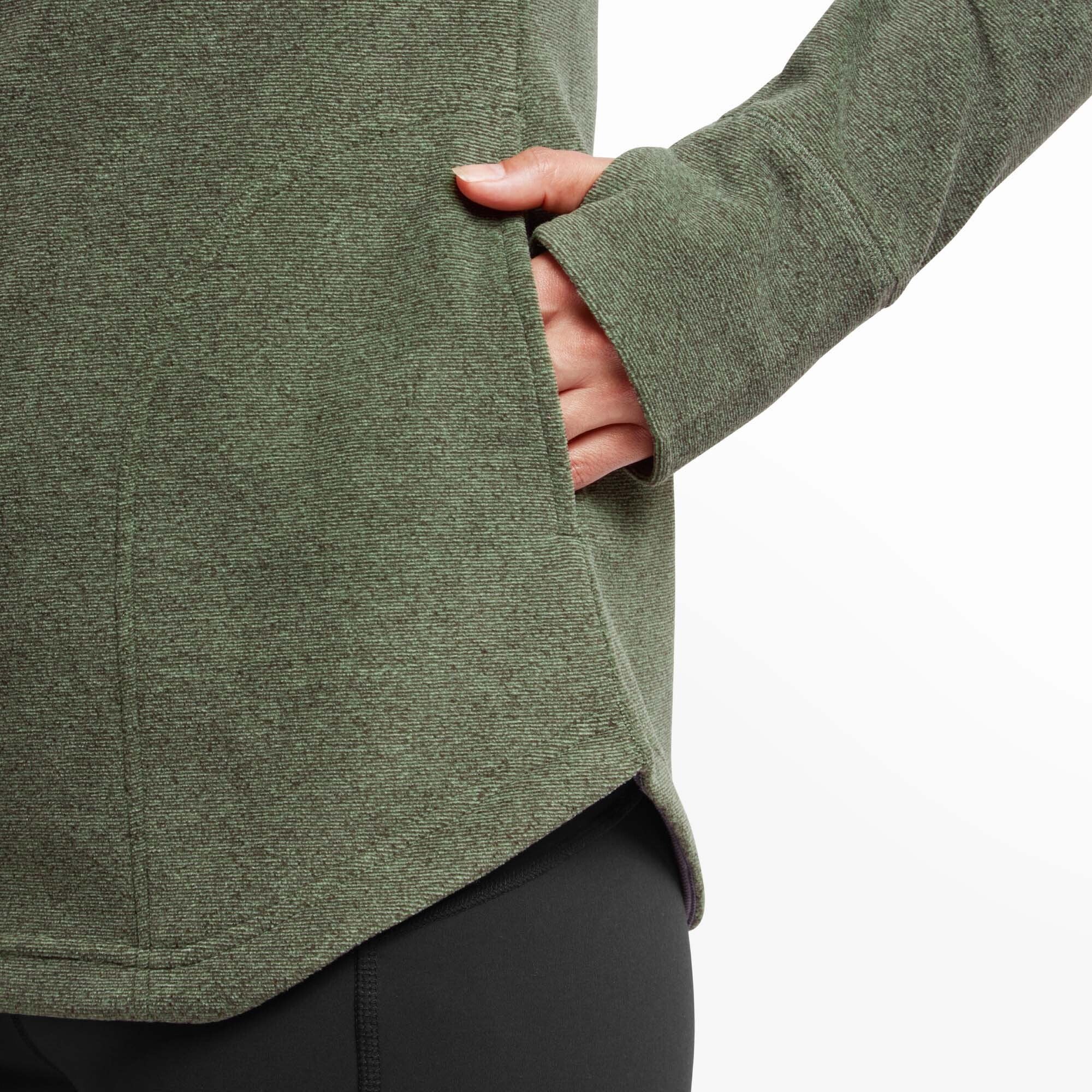 Close-up detail of the Sherpa Adventure Gear Rolpa Eco Pullover in Green's side pocket and thumbhole cuff. The model's hand rests inside the pocket, showcasing the convenient design, and the sleeve extends over her hand with a thumbhole opening for added warmth.