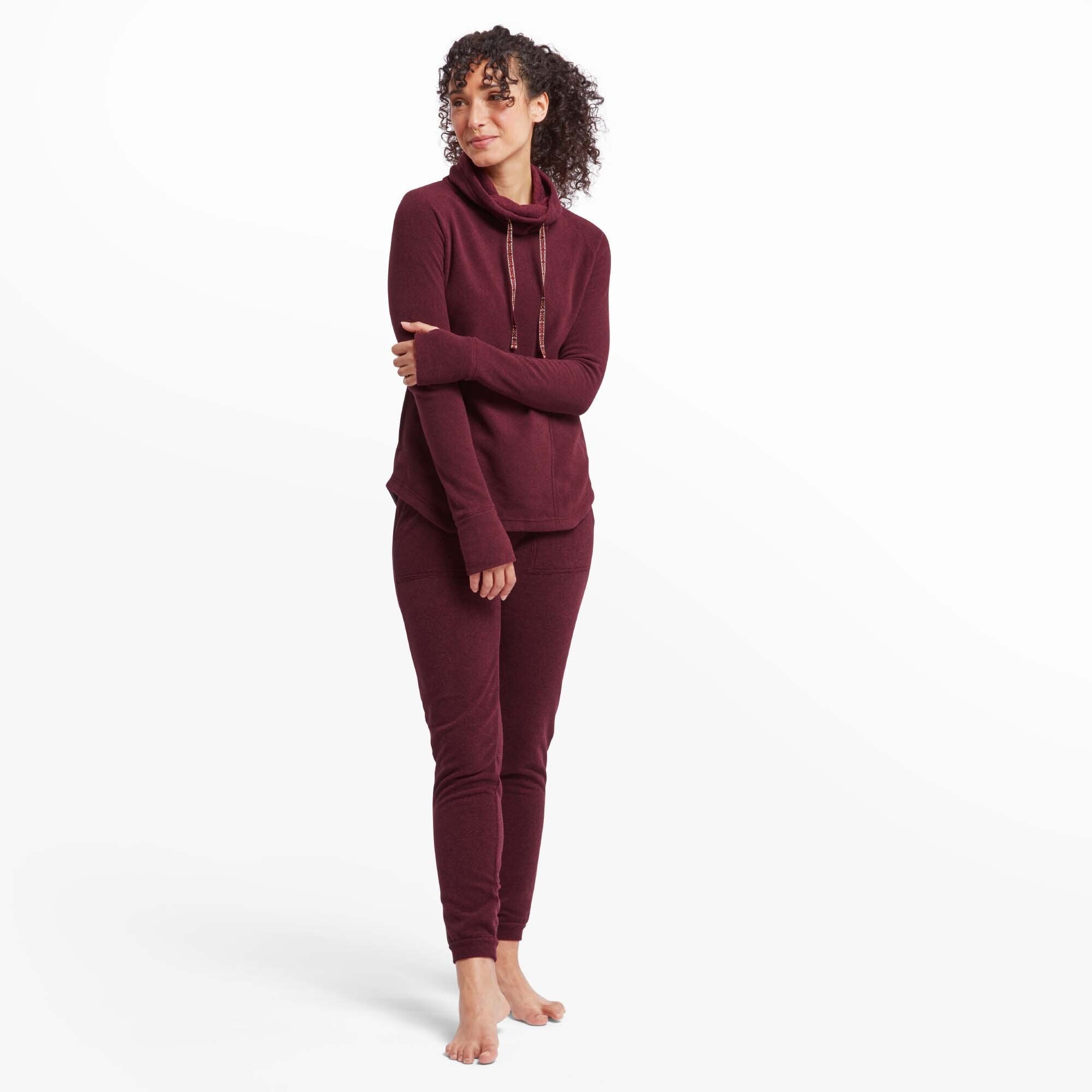 The same model stands barefoot in a full-body shot, wearing the Sherpa Adventure Gear Rolpa Eco Pullover in Red along with matching joggers. She has her arms crossed and smiles softly. The pullover’s relaxed fit and slightly curved hem are visible, as well as its soft, textured fabric.