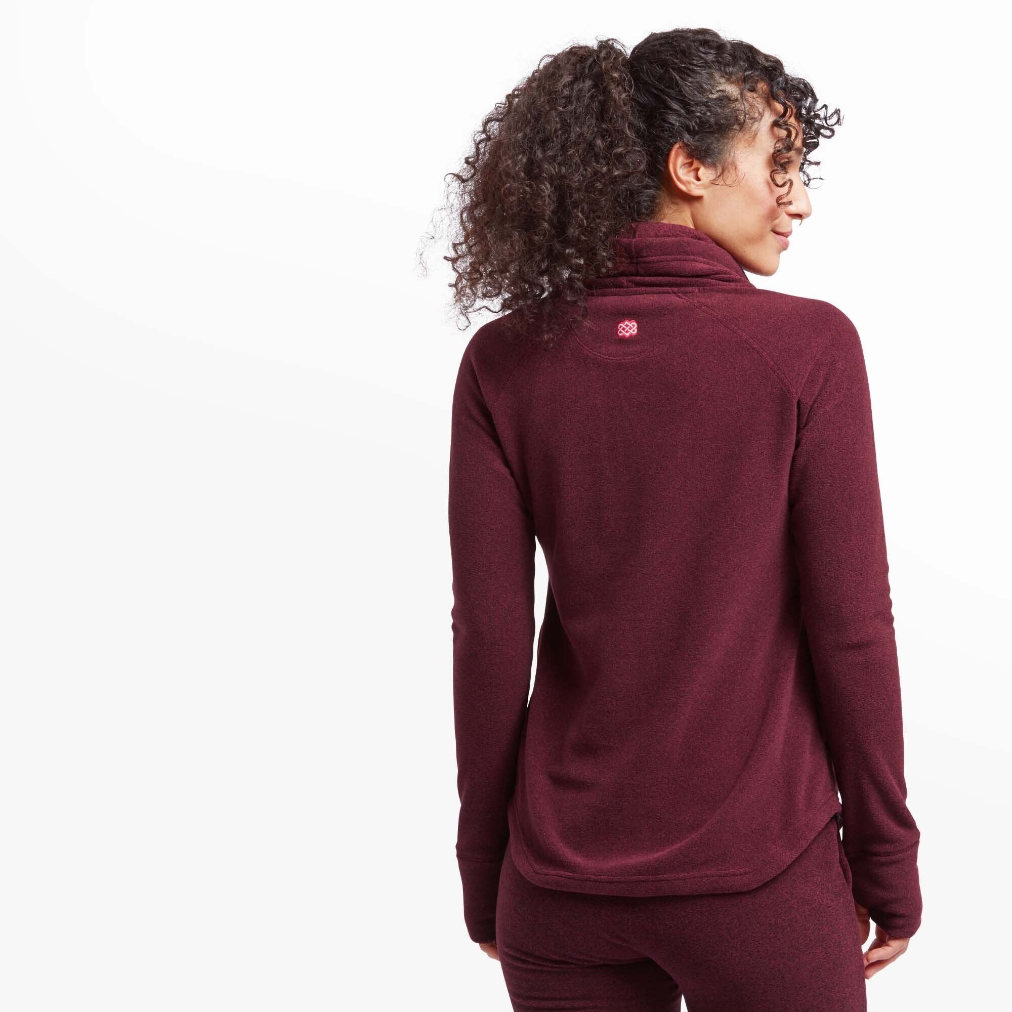 The model turns to show the back of the Sherpa Adventure Gear Rolpa Eco Pullover in Red, highlighting a small embroidered Sherpa logo centered just below the collar. The cowl neck drapes naturally at the back, and the curved hem provides coverage. Raglan sleeves contribute to the casual and comfortable fit.