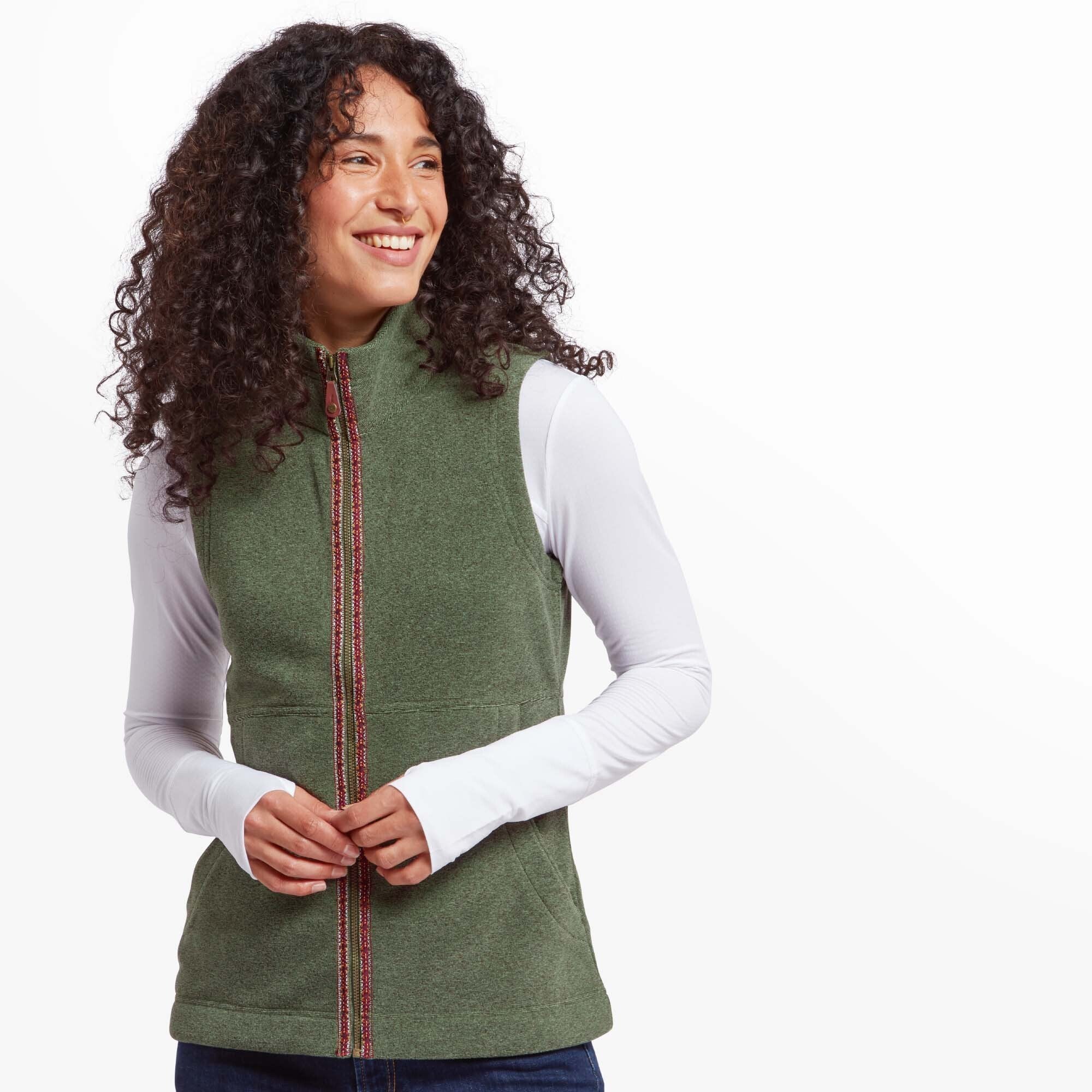 A woman is wearing a Sherpa Adventure Gear Rolpa Eco Vest in Green over a white long-sleeve shirt. The vest features a straight fit with a high neckline and decorative embroidered trim running along the zippered front. She is smiling and looking to the side, with her hands lightly clasped in front.