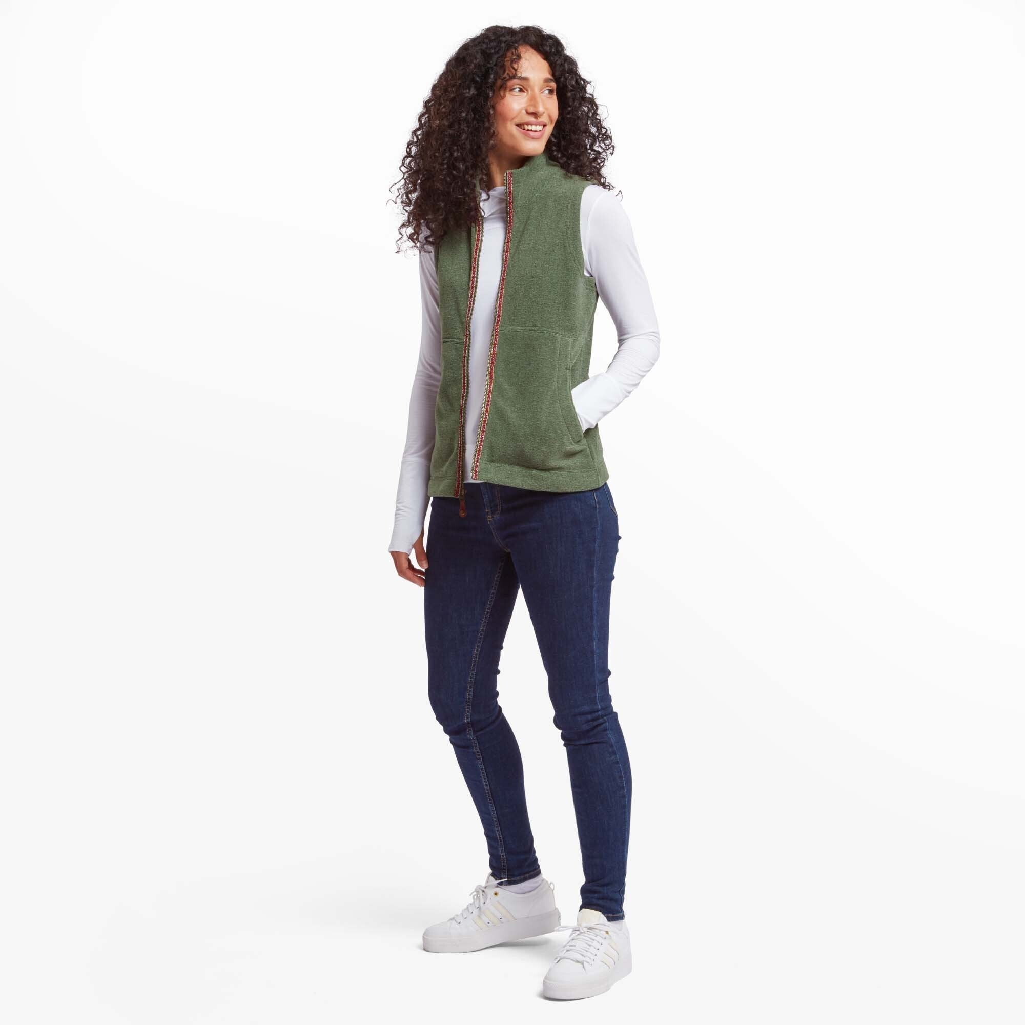 The same woman is seen in a full-body shot, showcasing the Sherpa Adventure Gear Rolpa Eco Vest in Green paired with dark blue jeans and white trainers. She stands confidently, with one hand in her pocket and a cheerful expression, looking slightly to the side.