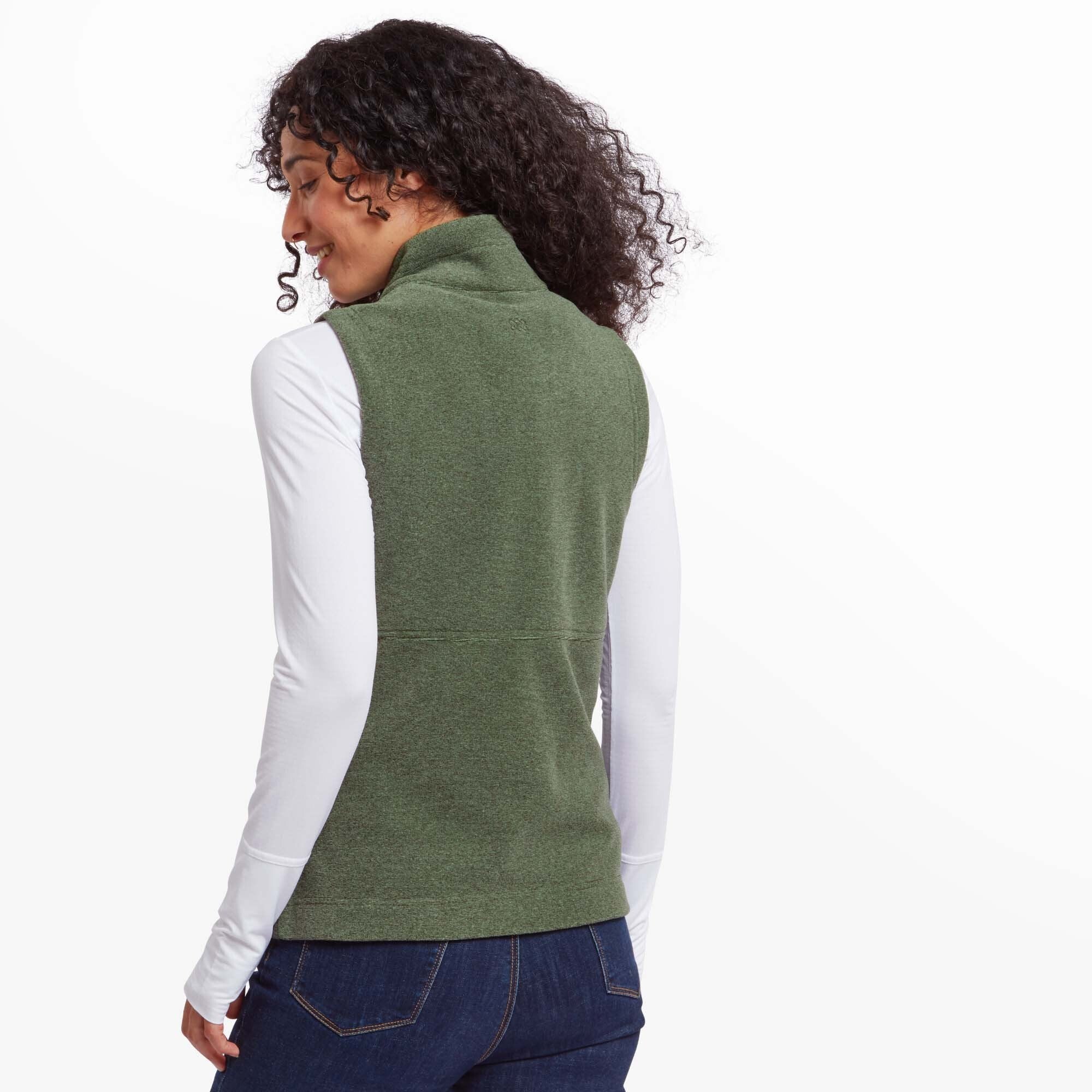 The back view of the Sherpa Adventure Gear Rolpa Eco Vest in Green, highlighting its clean, seamless design and the high neckline. The woman’s curly hair is visible, and she is wearing the same white long-sleeve shirt and jeans.