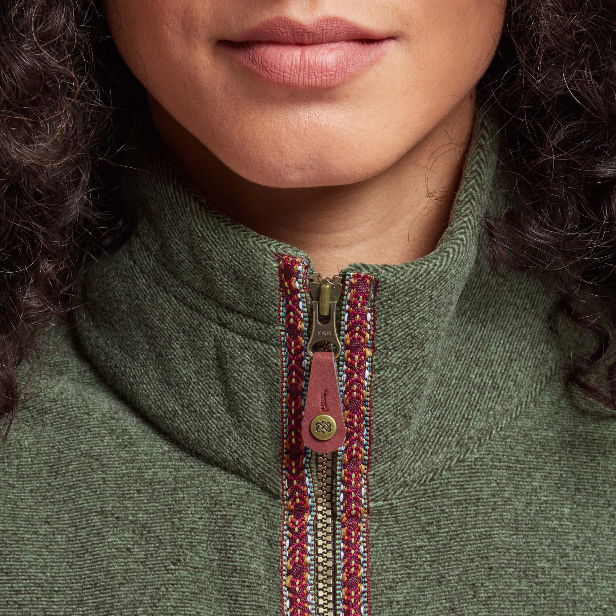 A close-up of the Sherpa Adventure Gear Rolpa Eco Vest in Green’s zipper, showing intricate red and multicolored embroidered detailing along the trim and a brass zipper pull with leather detailing. The texture of the vest fabric is visible, adding depth to the image.