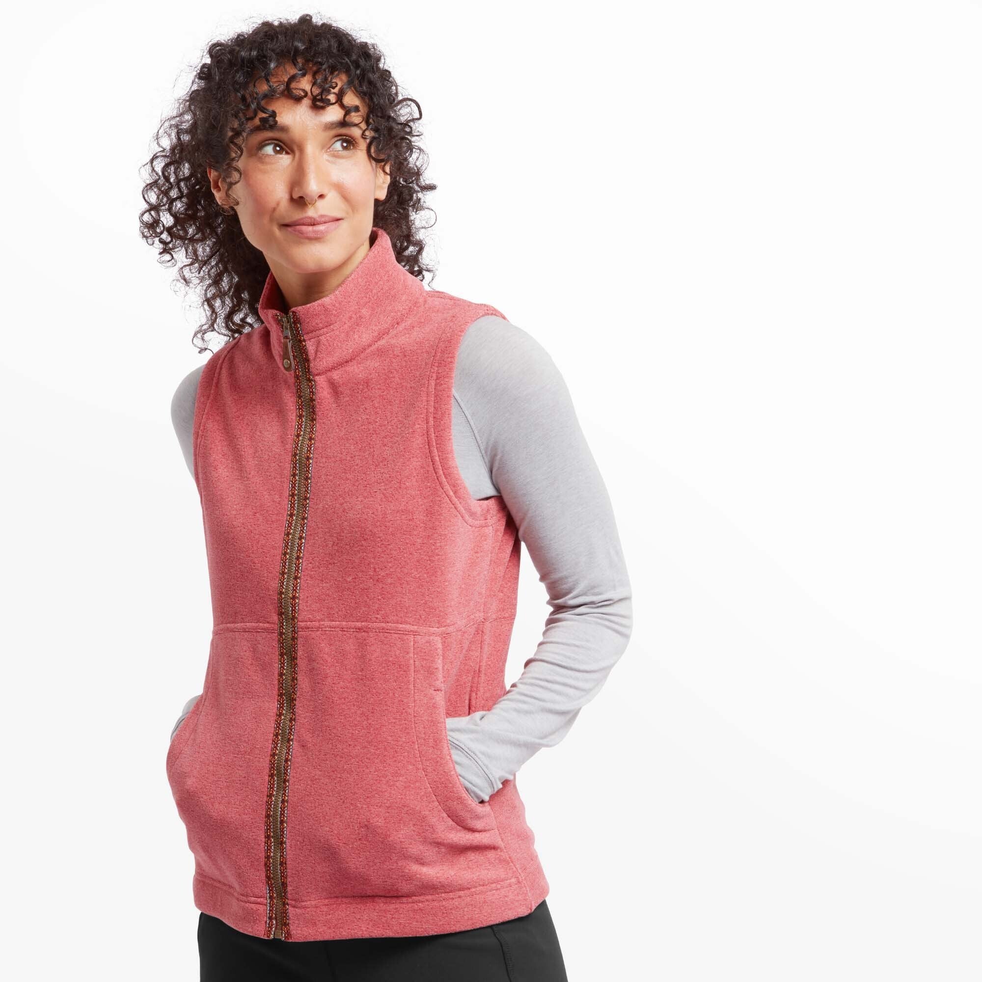 A model wearing a Sherpa Adventure Gear Rolpa Eco Vest in Red with a high collar, paired with a long-sleeved light grey top. The vest features a full zip closure with intricate multicolor embroidery along the zipper line, adding a decorative touch. The model has her hands in the side pockets, standing in a relaxed pose against a light background.