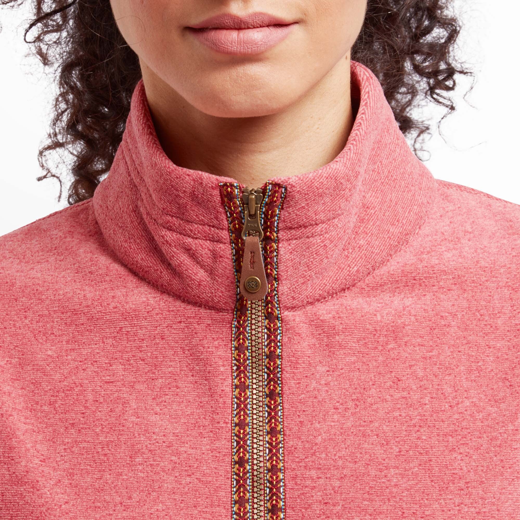 A close-up of the Sherpa Adventure Gear Rolpa Eco Vest in Red's high collar and zipper details. The multicolor embroidered trim along the zipper and the leather zipper pull are clearly visible, showcasing the craftsmanship and attention to detail in the design. The coral-pink fabric texture is soft and cozy, enhancing the overall aesthetic.