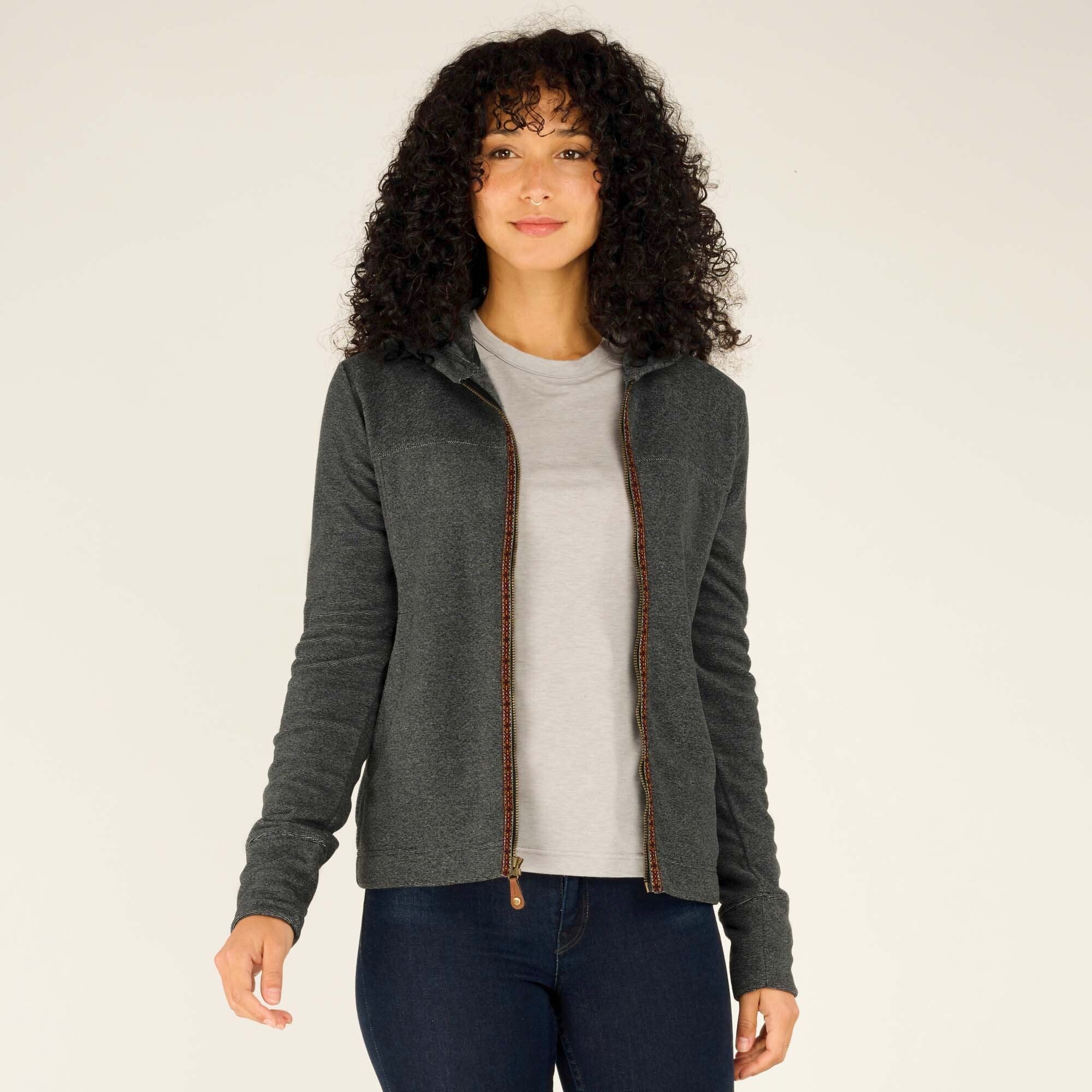 A female model with curly hair wears a Sherpa Adventure Gear Rolpa Full Zip Hoodie in Grey. The hoodie features a subtle texture, a full-length front zipper with a decorative woven trim, and a comfortable fit. She pairs the hoodie with a light grey T-shirt and dark blue jeans, standing against a plain off-white background.